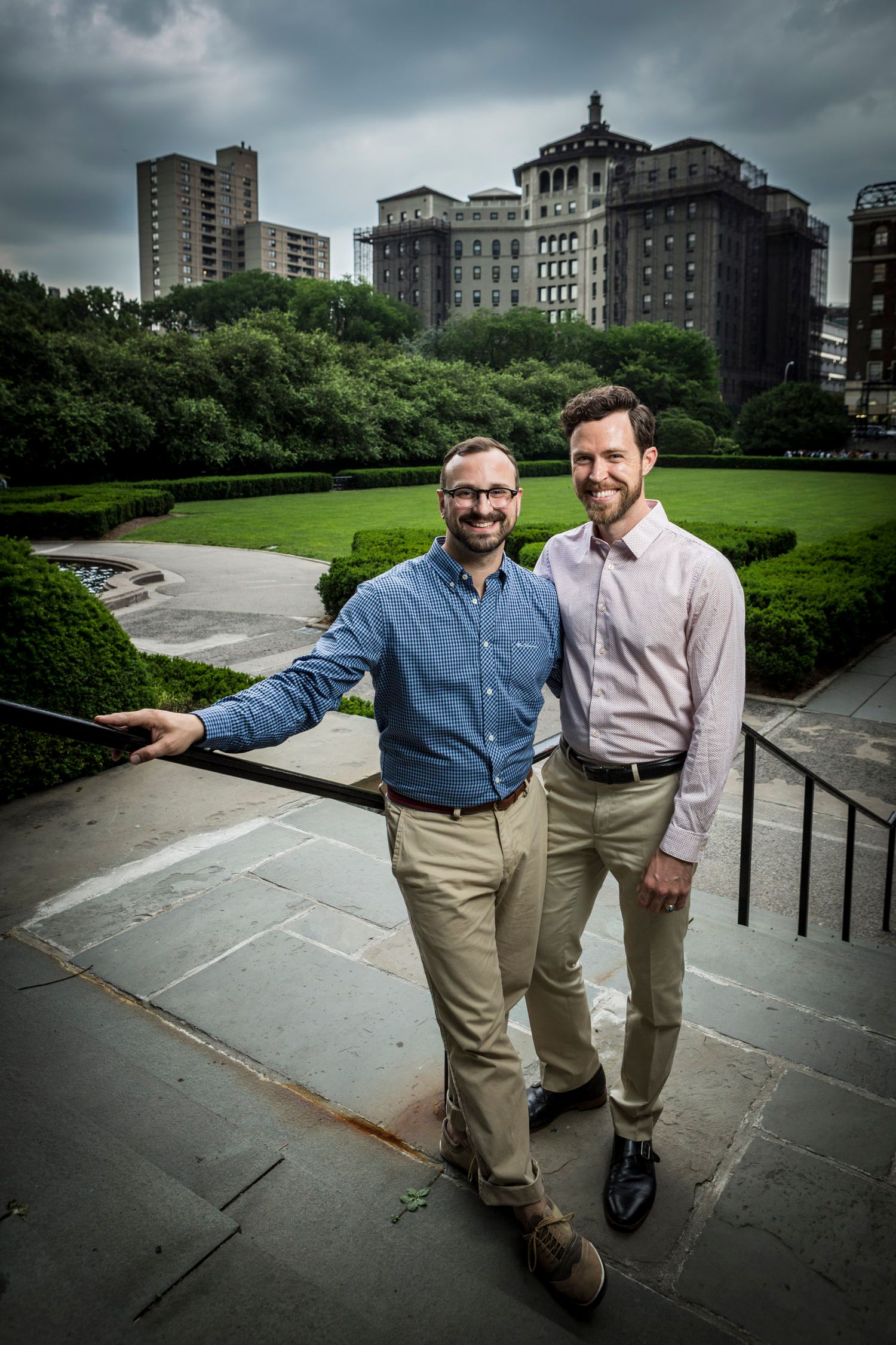 Two smiling men pose together on stone steps with lush green park and city buildings in the background.