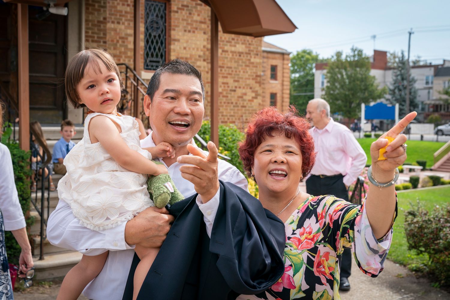 Asian man holding toddler girl in white dress with older woman pointing outdoors near brick church.
