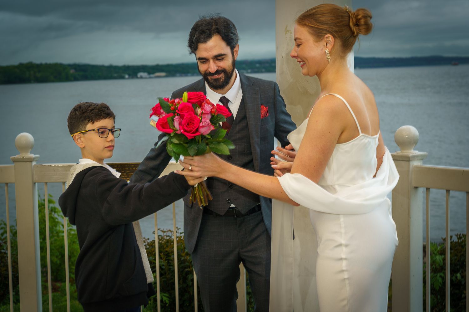A young boy presents red roses to a bride in a white dress and groom in gray suit at an outdoor waterfront wedding.