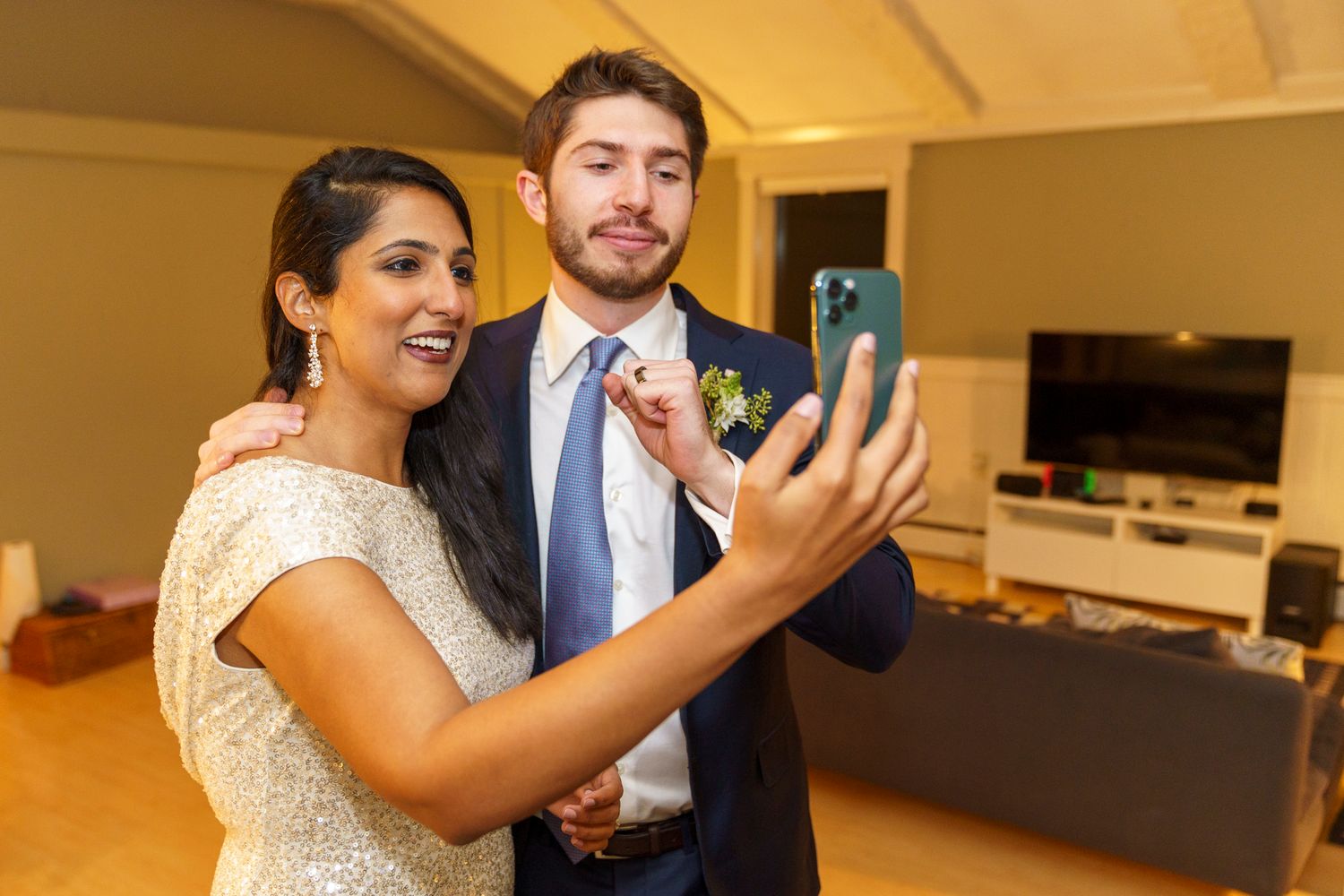 Couple in formal attire takes a selfie together, man in suit with boutonniere, woman in sparkly dress, indoors.