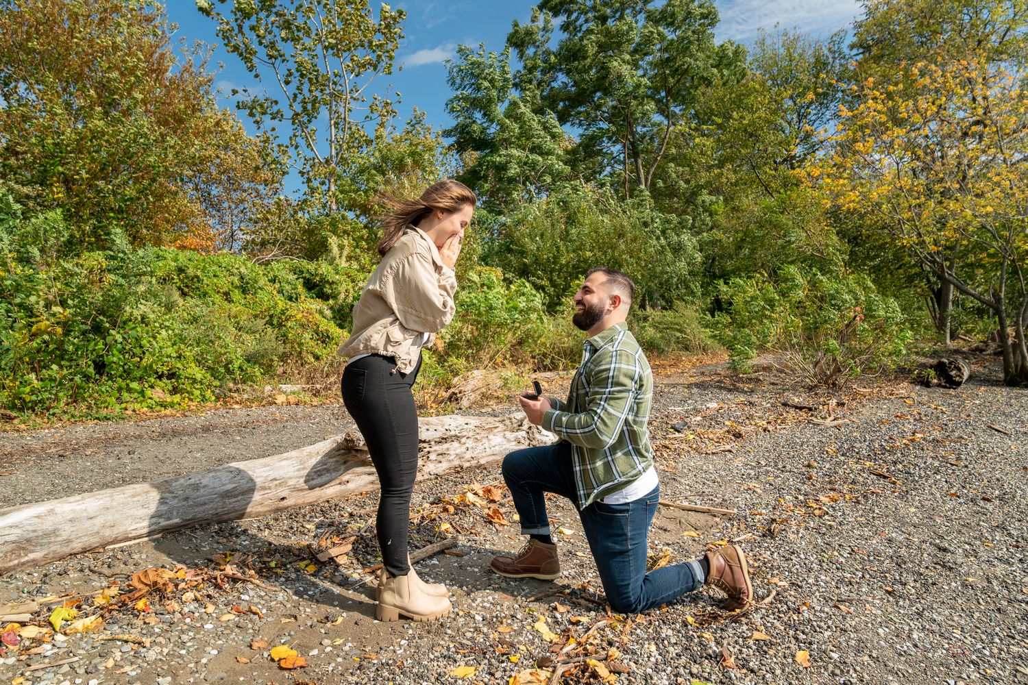 Man proposes on one knee to woman outdoors on gravel path surrounded by autumn foliage on sunny day.
