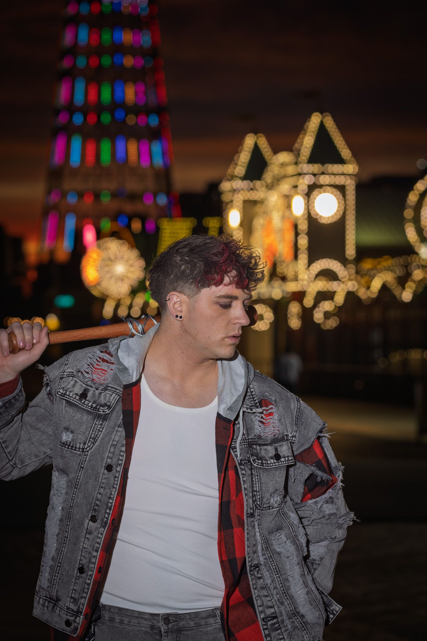 Young man in a distressed denim jacket and plaid shirt holds a bat on his shoulder in front of glowing carnival lights at night.