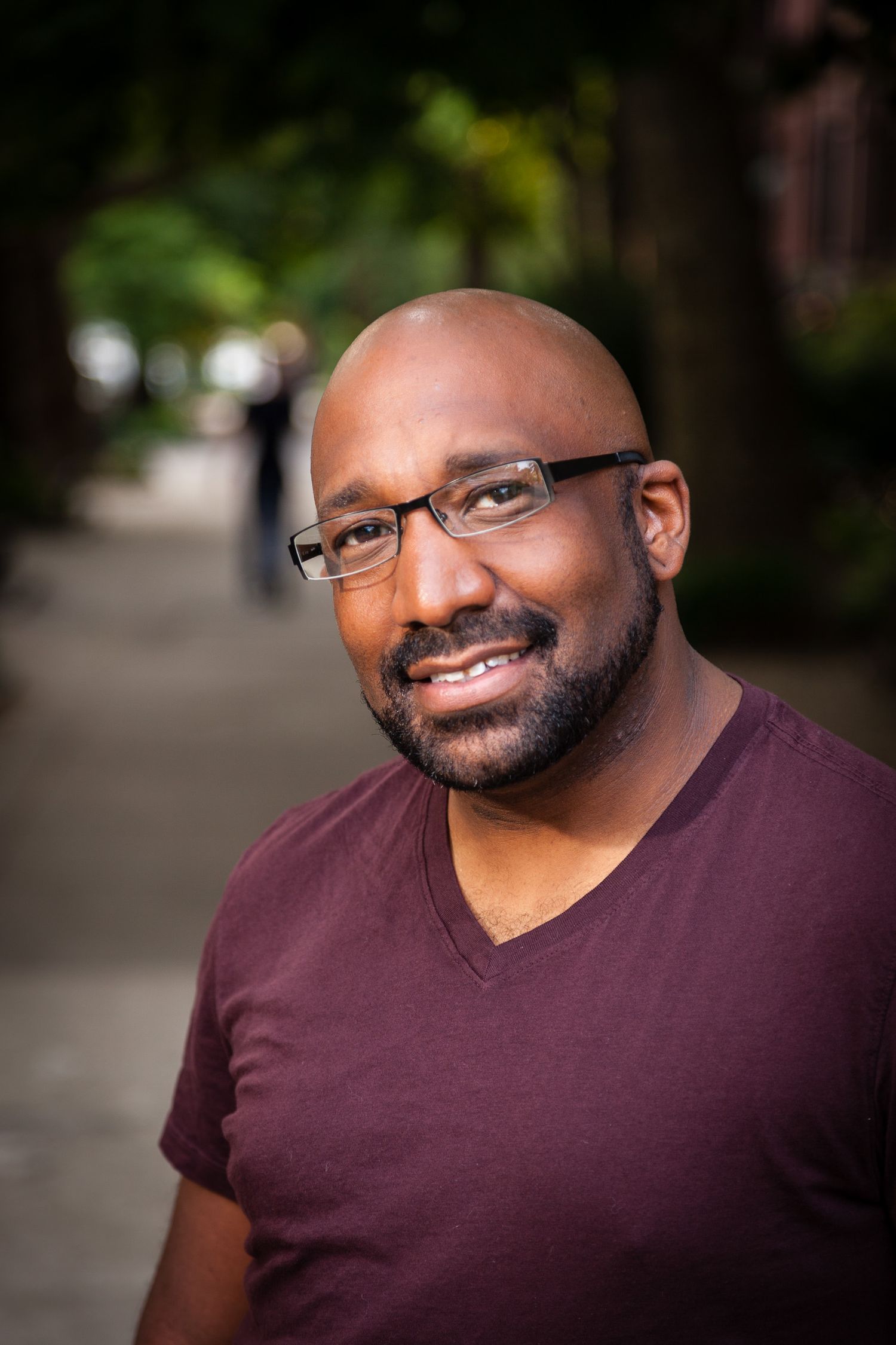 Bald Black man with glasses and beard smiling outdoors on tree-lined sidewalk.
