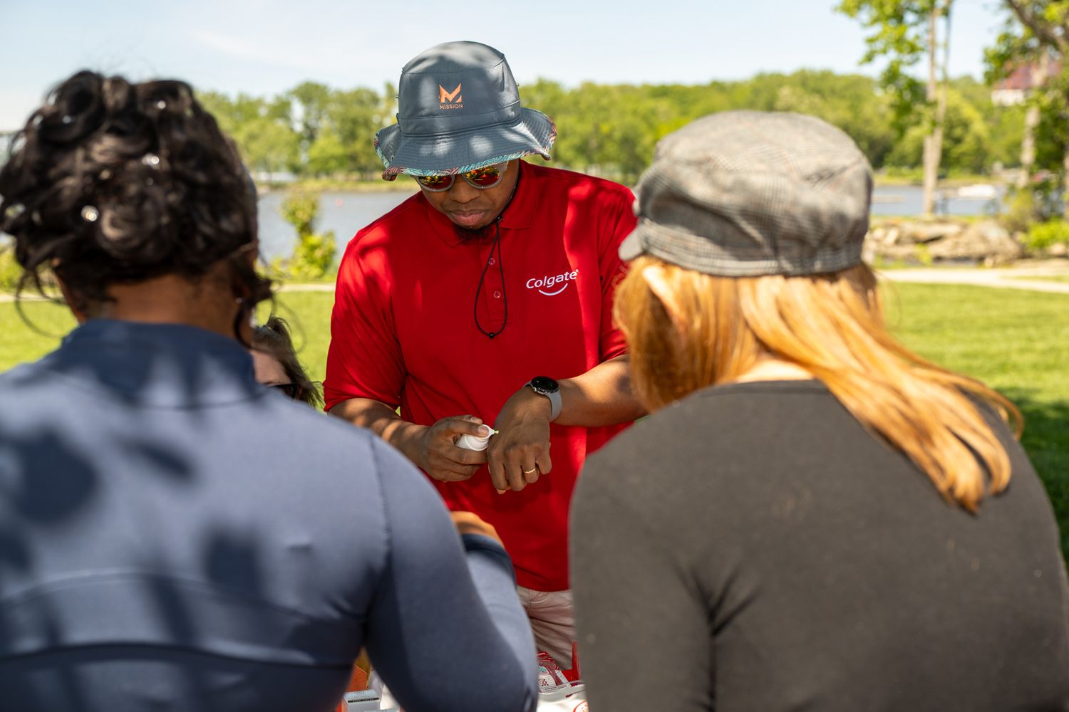 A man in a red shirt and bucket hat shows something to two people outdoors in a sunny park setting.