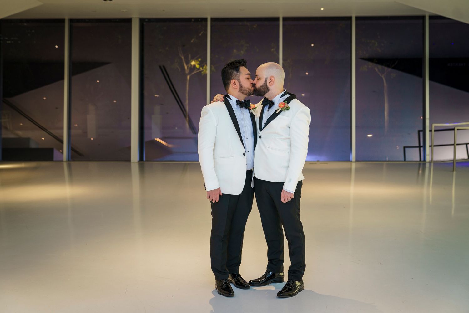 Two grooms in white tuxedos sharing a kiss on dance floor during their wedding ceremony.