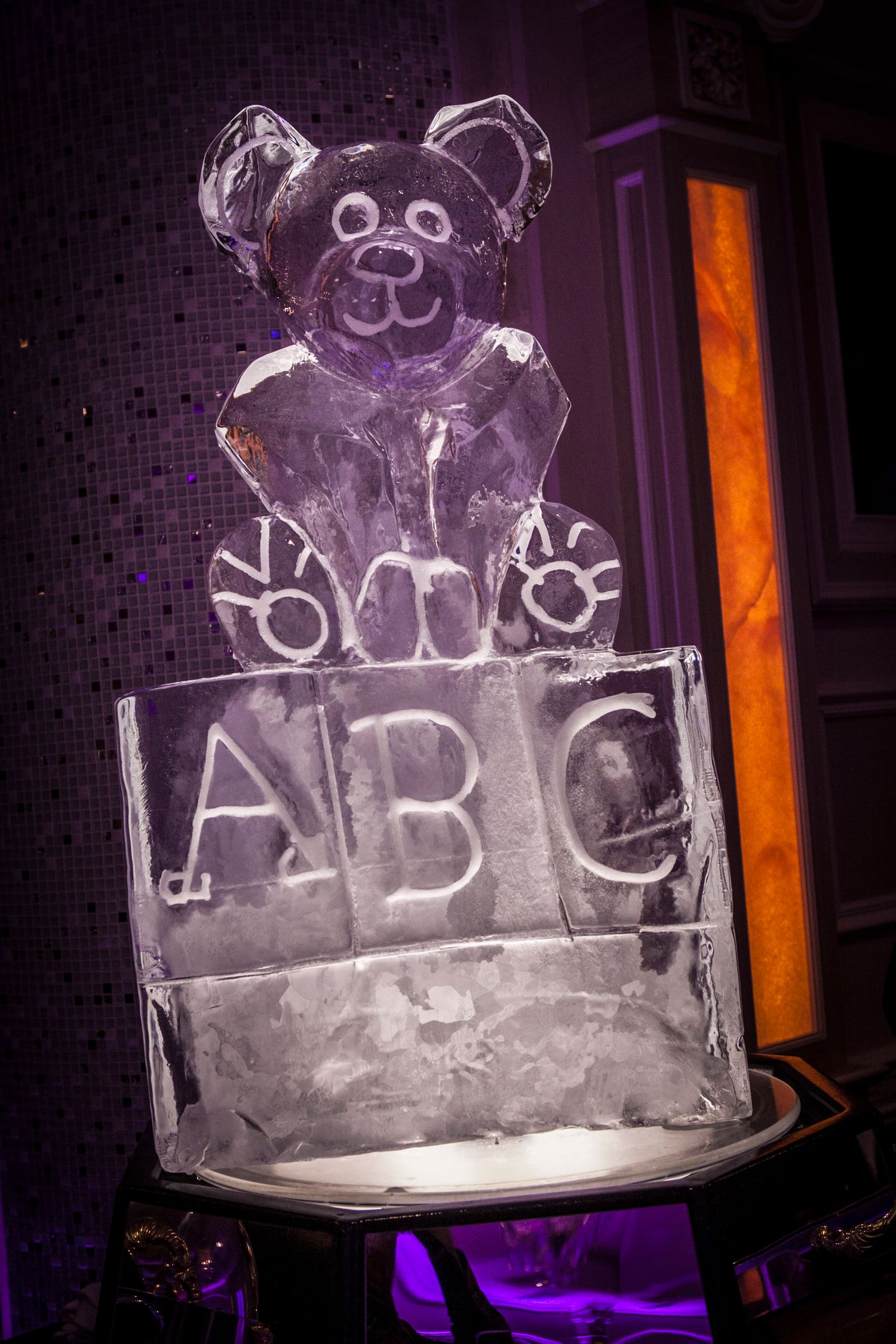 An intricate ice sculpture of a teddy bear sitting on ABC blocks at a baby shower event.