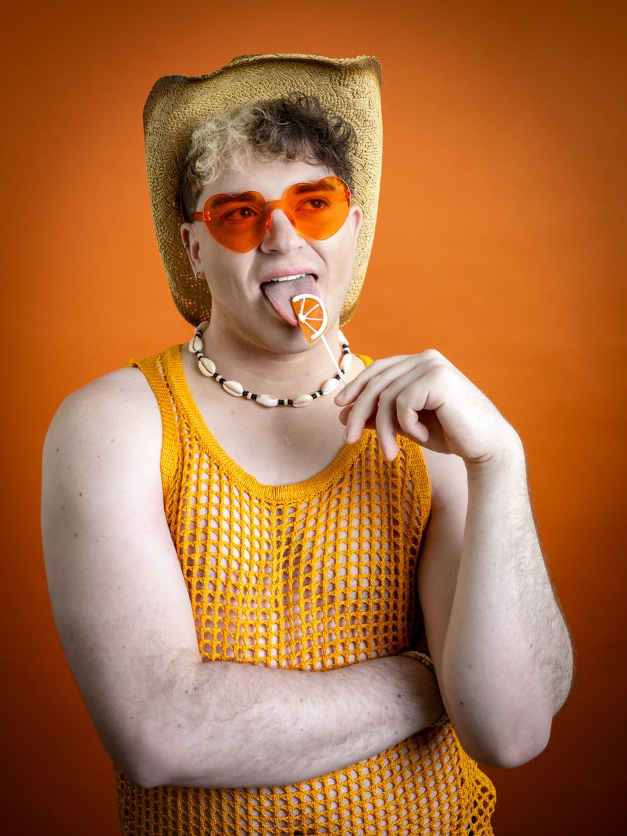 Man in orange mesh top and orange heart sunglasses licking a lollipop against an orange background.