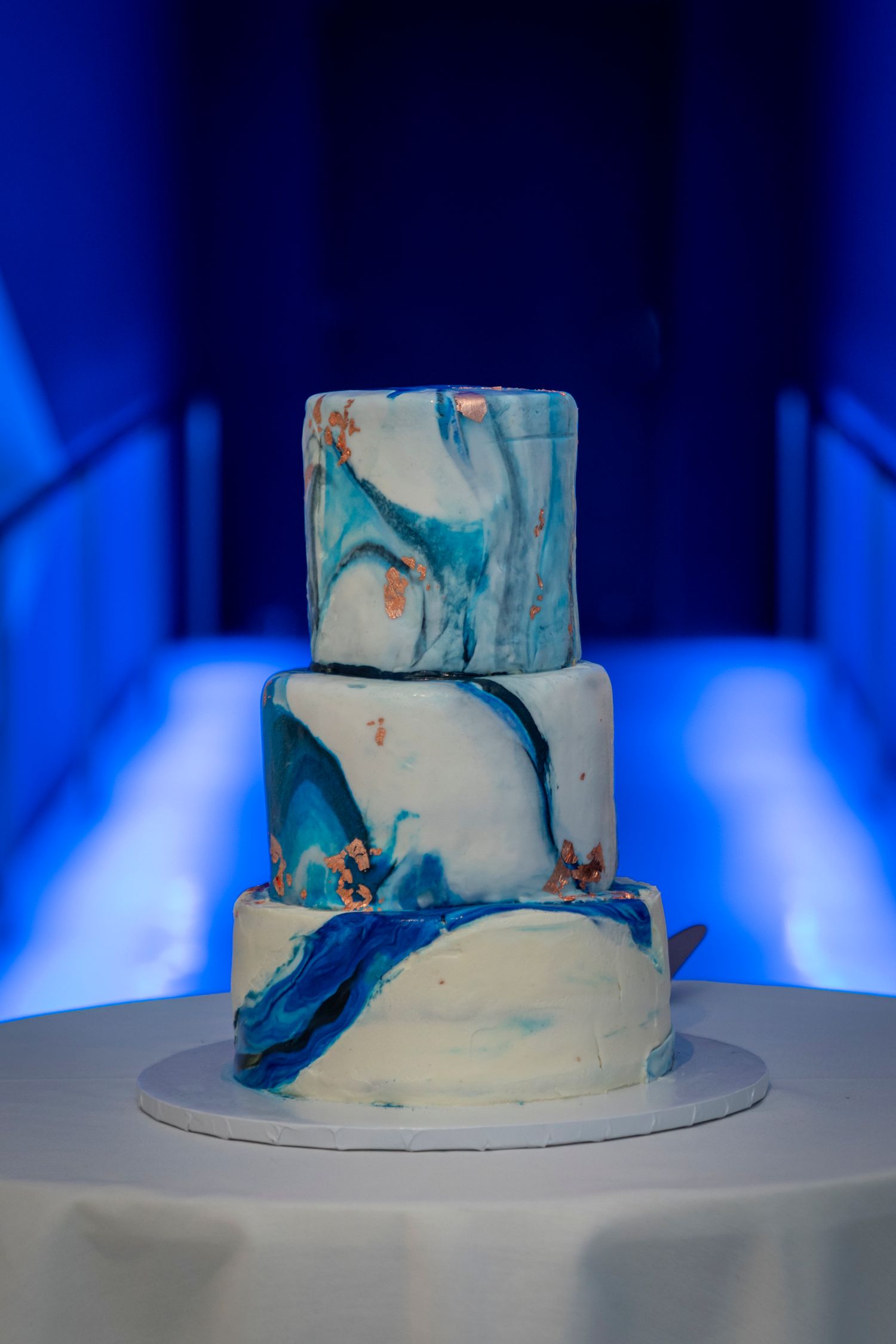 Three-tier marble fondant wedding cake with blue and white swirls and gold accents on a white stand.