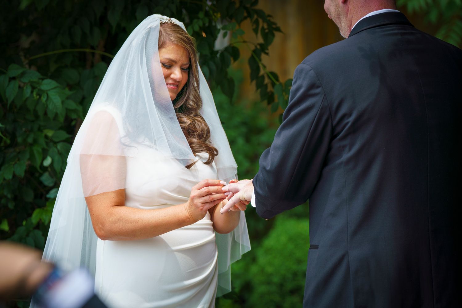 Bride in white veil placing wedding ring on groom's finger during outdoor ceremony with lush green foliage backdrop.