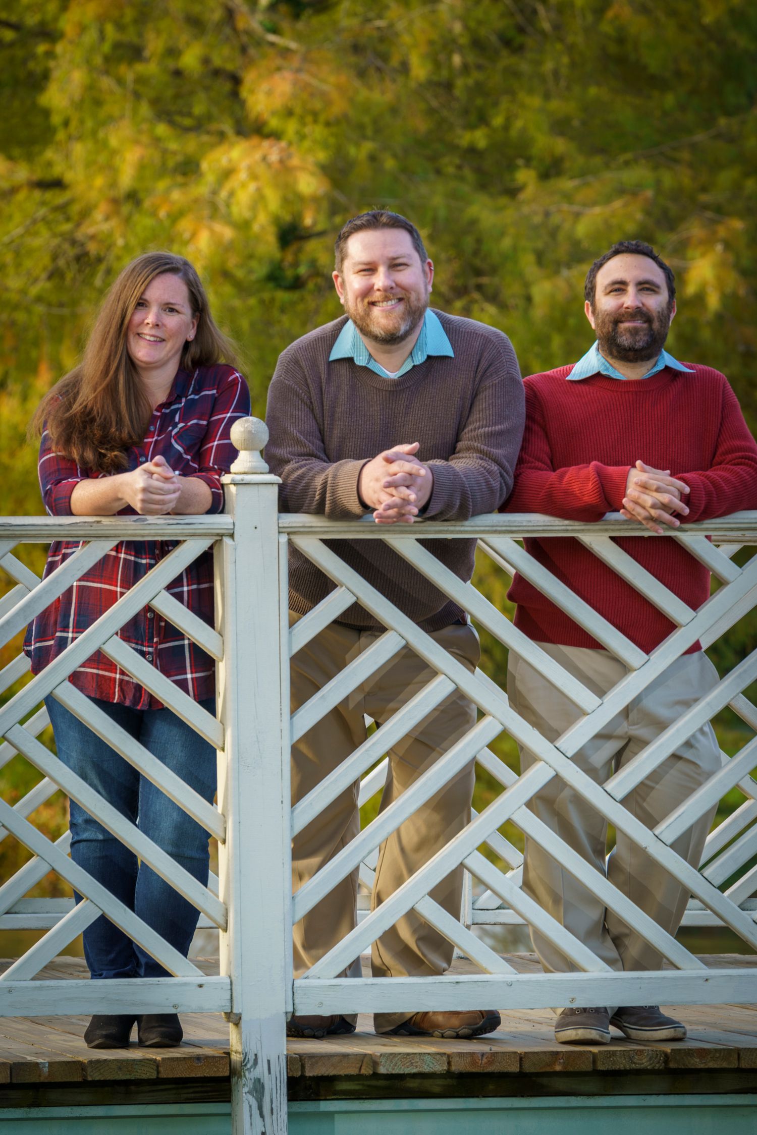 Three smiling friends leaning on a white decorative bridge railing outdoors in autumn.