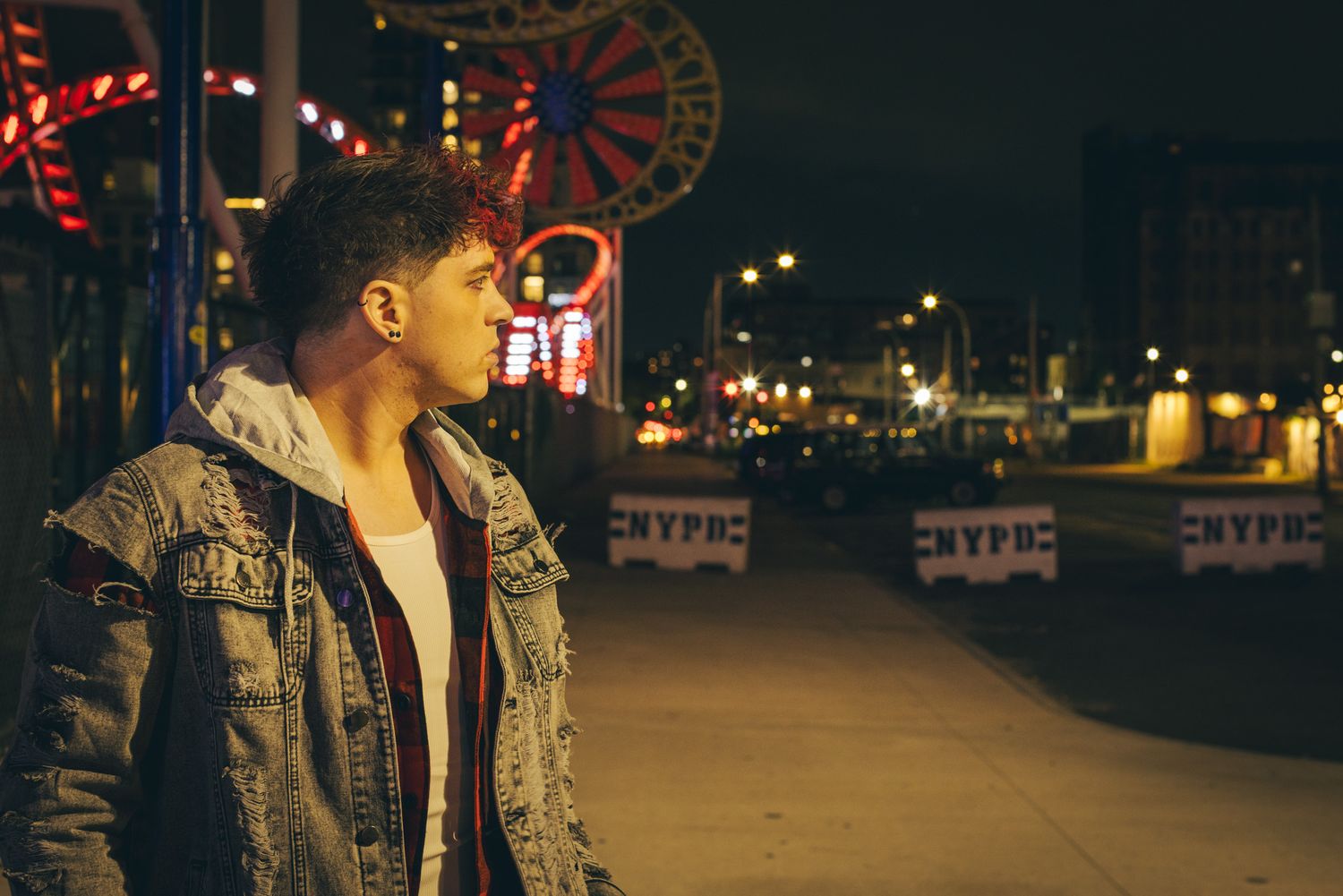 Young man in a denim jacket and plaid shirt gazes sideways at night near a boardwalk with NYPD barriers and a glowing carnival ride.