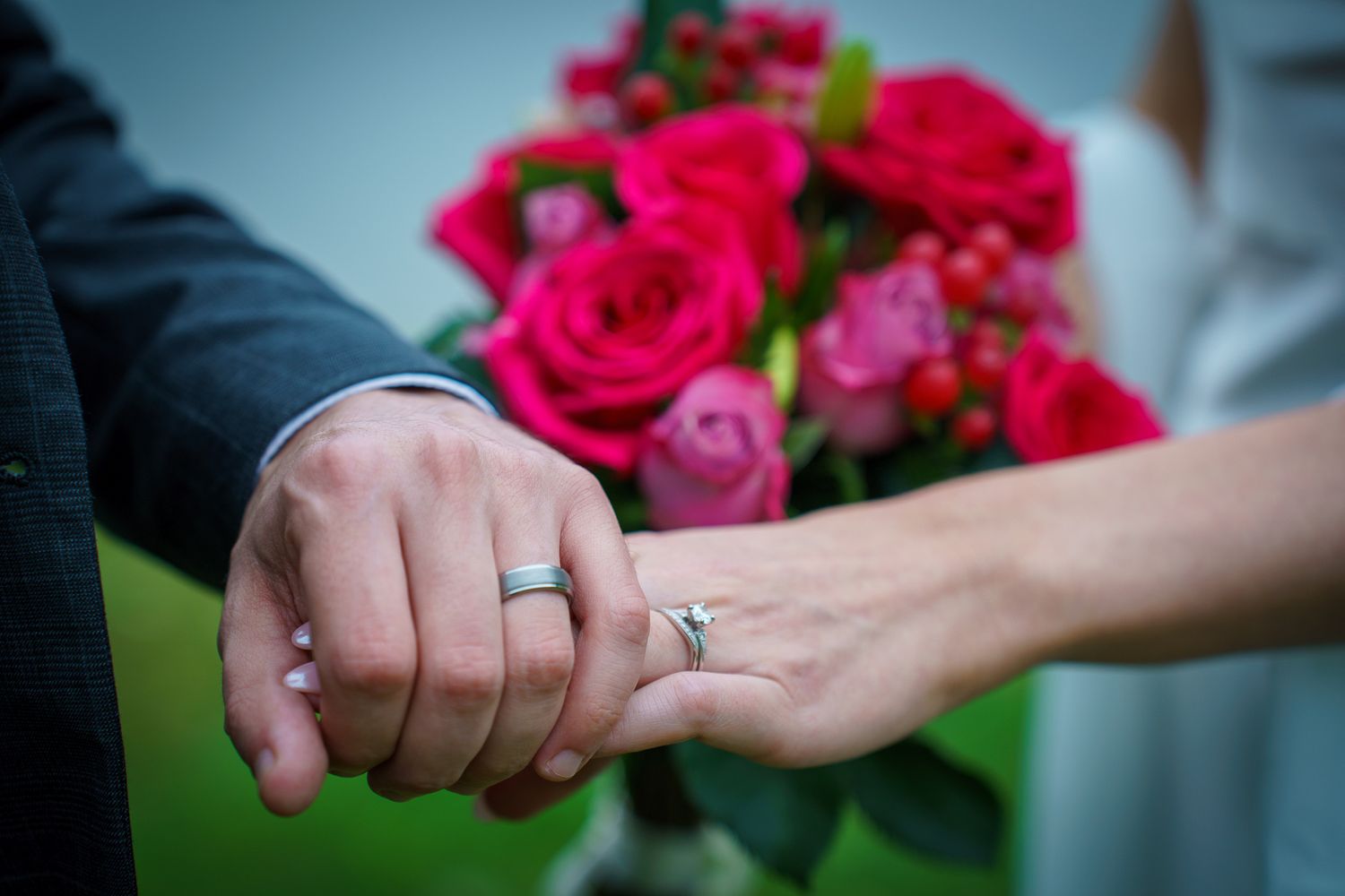 Newlywed couple holding hands showing wedding rings with a vibrant pink rose bouquet in the background.