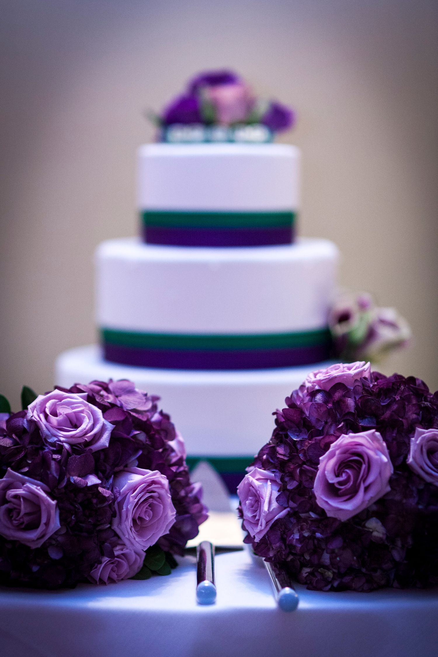 Three-tier white wedding cake with purple and green ribbon, flanked by two purple rose and hydrangea bouquets.
