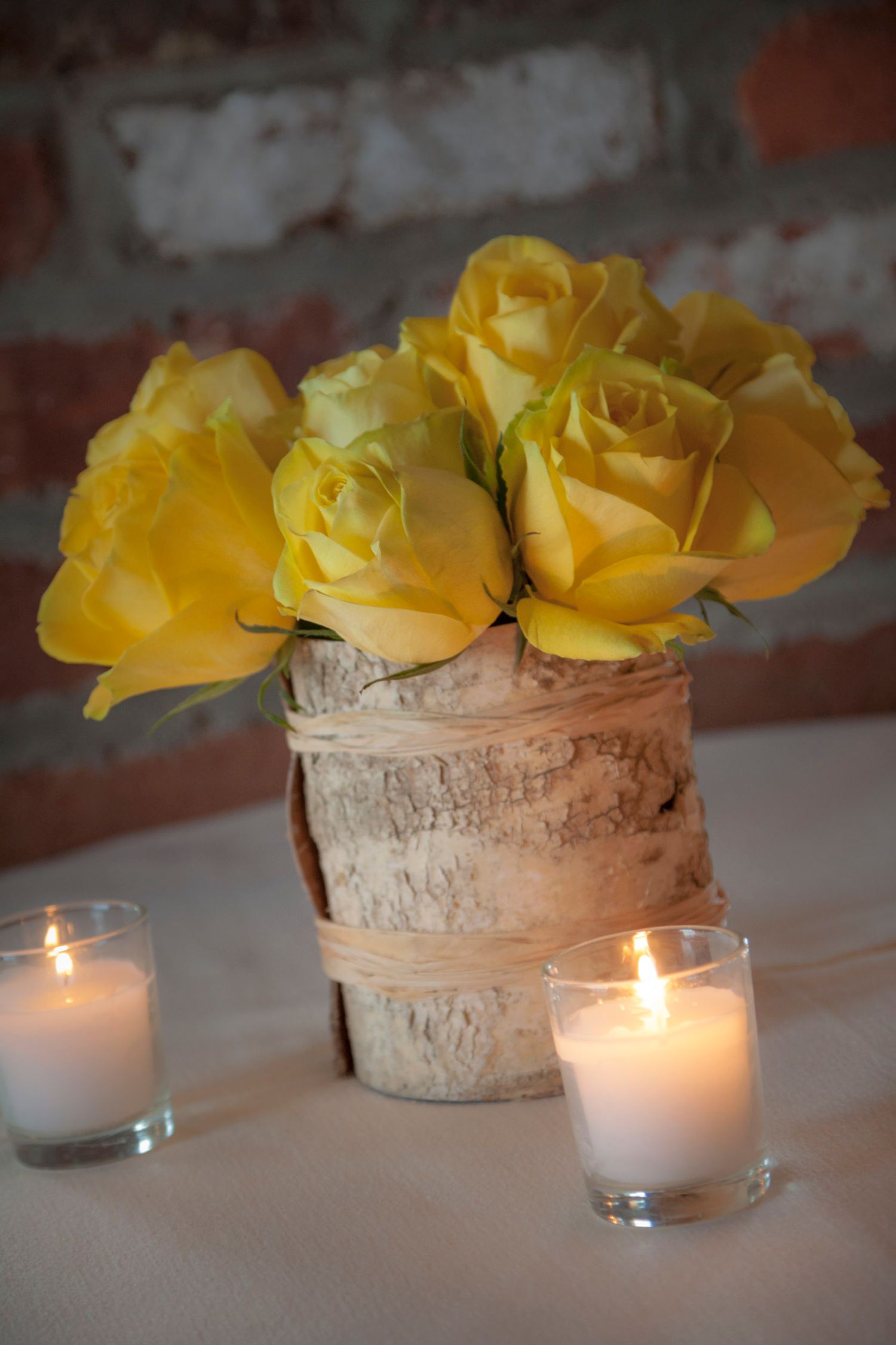 Yellow roses in a birch bark vase centerpiece flanked by lit candles on a white tablecloth.