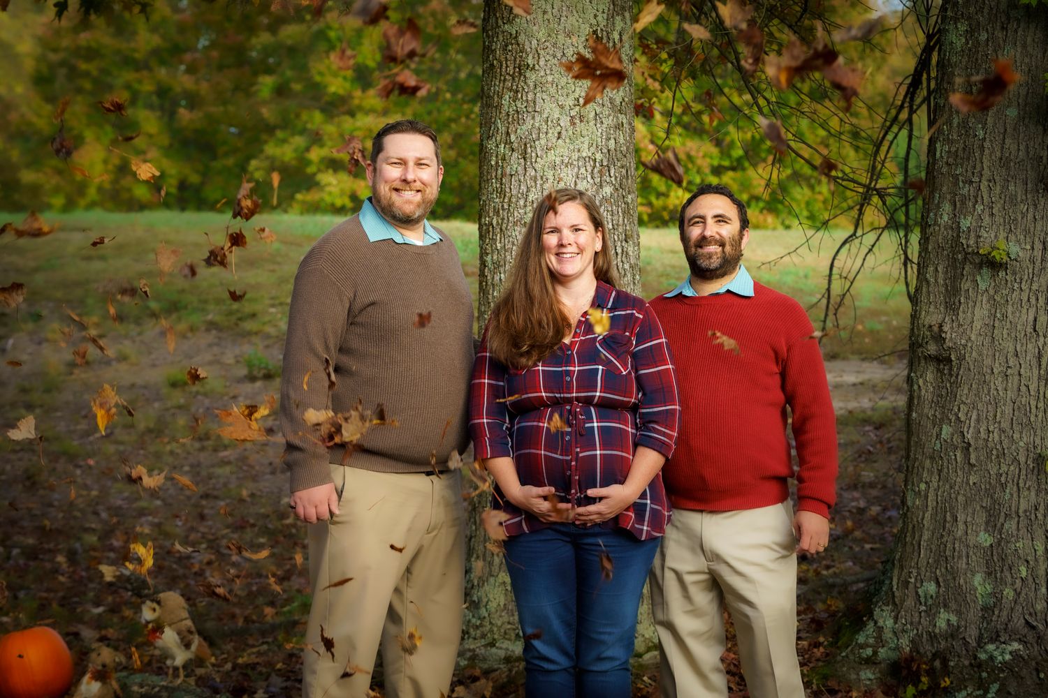 Three people smiling outdoors in autumn, standing near a tree with colorful fall foliage and a pumpkin nearby.