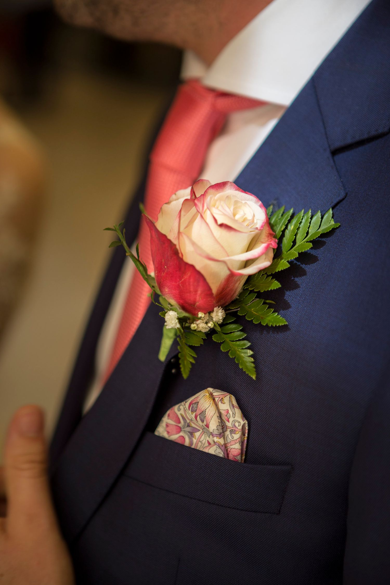 Man in navy suit with pink rose boutonniere, coral tie, and paisley pocket square at a formal event.