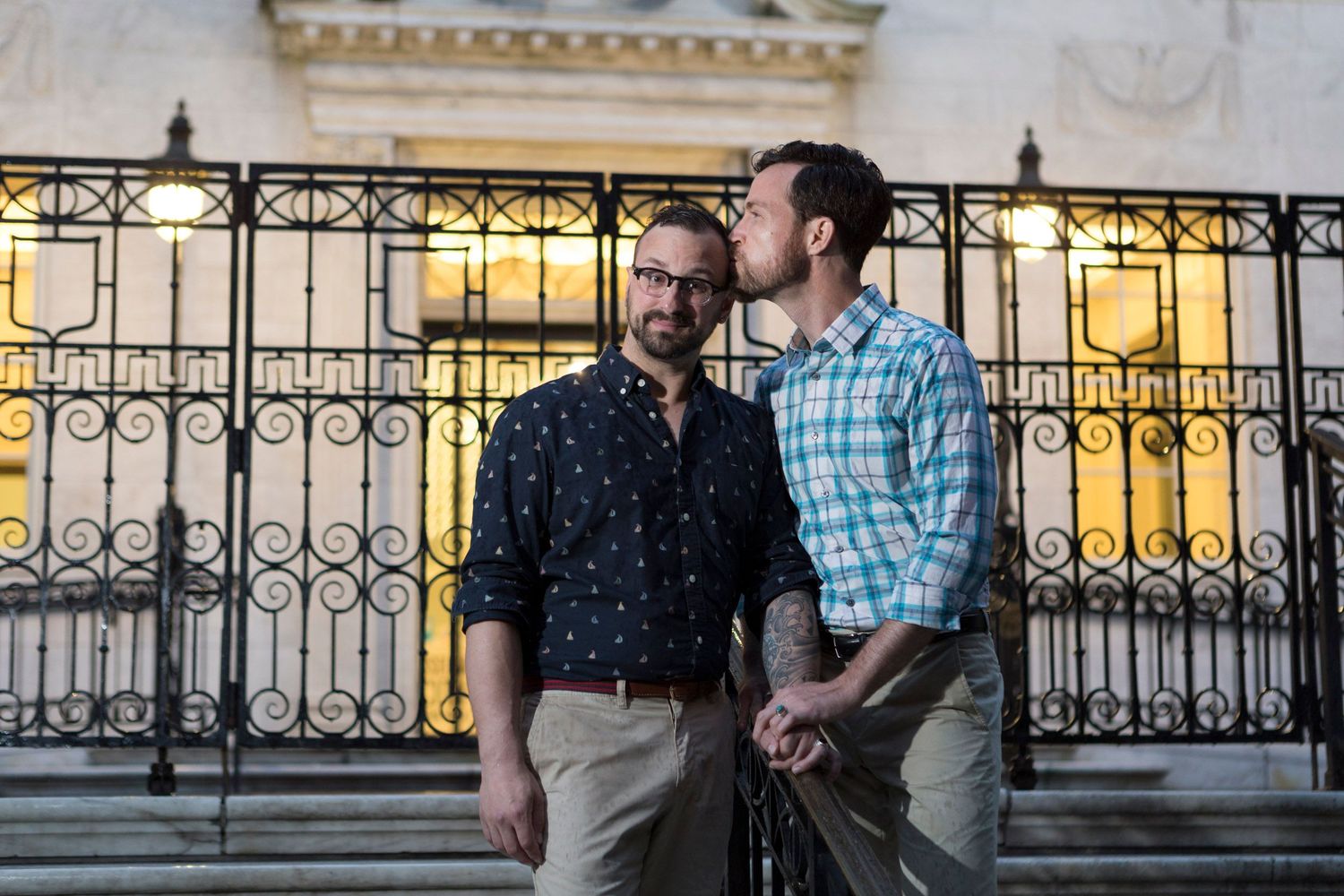 Two men share a tender moment on steps in front of an ornate iron gate at dusk.