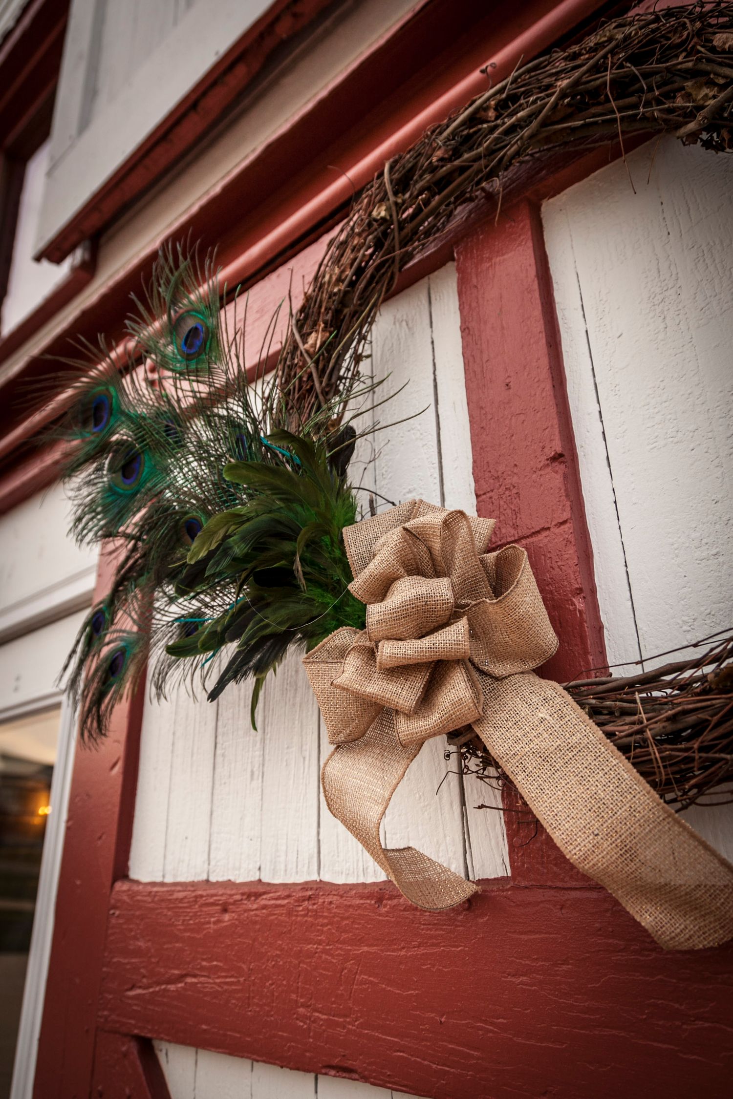 Rustic wedding wreath with peacock feathers and burlap bow on red barn door.