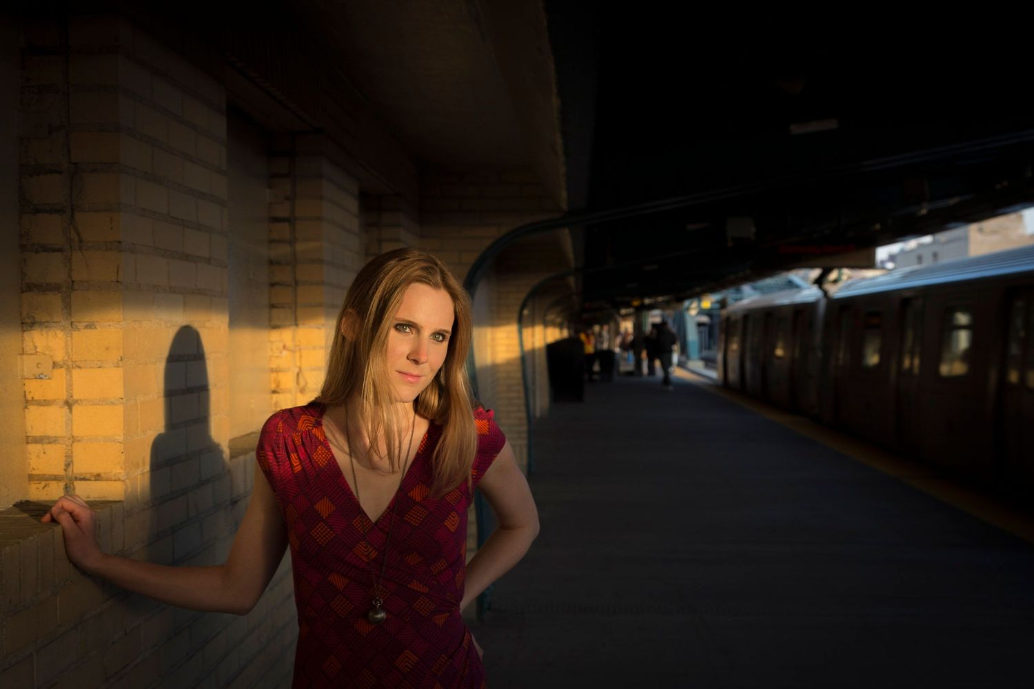 Woman in red dress standing on subway platform at golden hour, with train and dramatic shadows behind her.