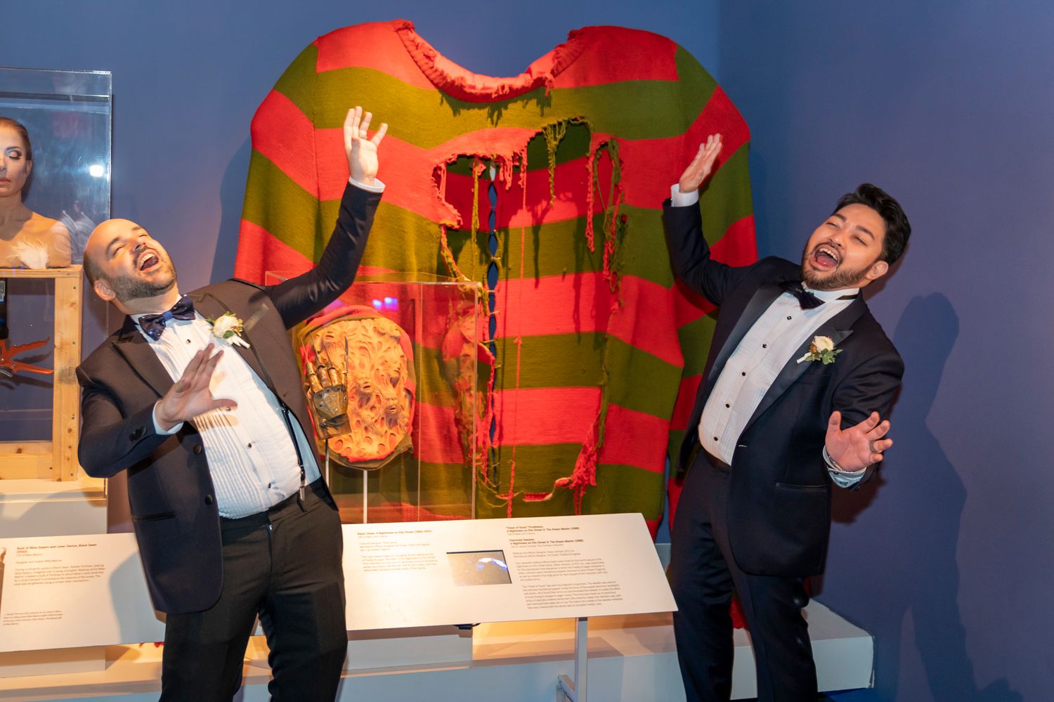 Two men in tuxedos pose joyfully in front of a large Freddy Krueger sweater artwork at a museum exhibit.