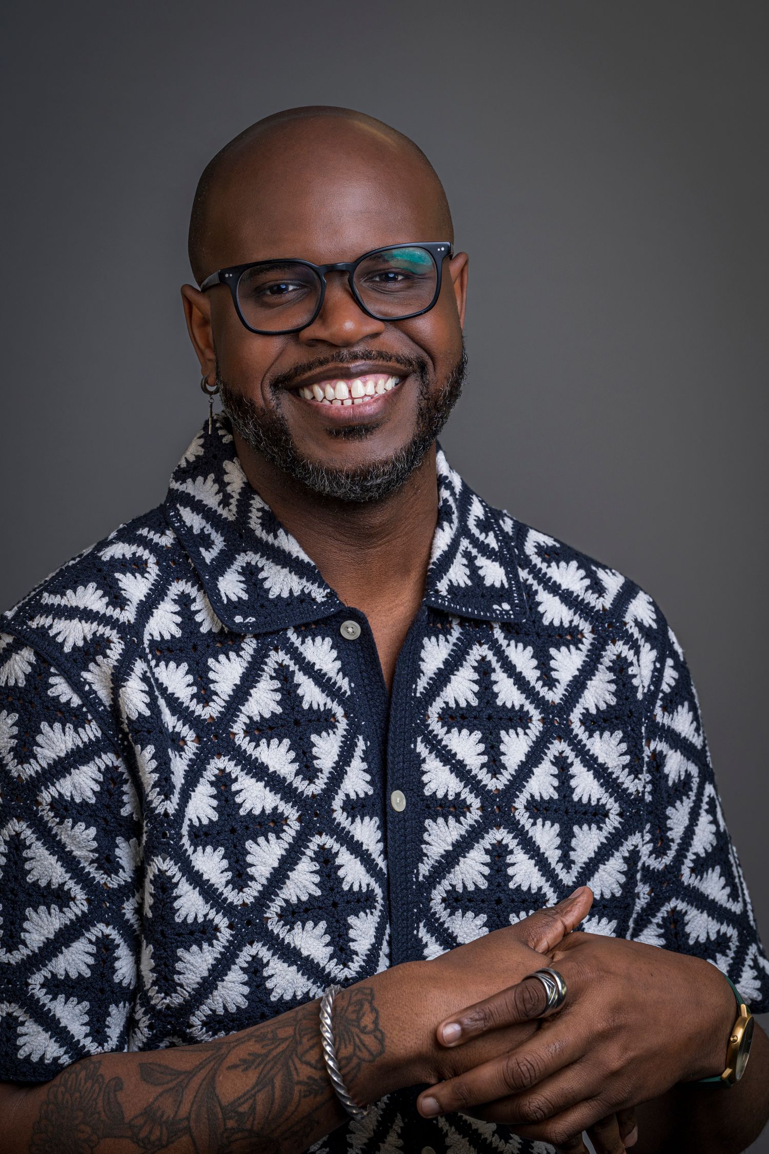 Smiling bald Black man with glasses, beard, tattoos, wearing a patterned blue and white shirt, arms crossed.