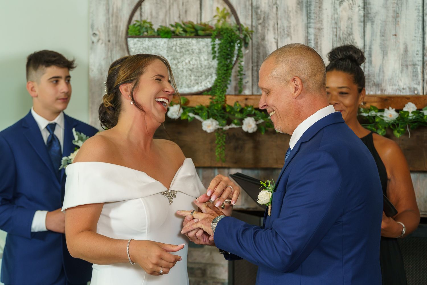 Bride and groom exchange rings at wedding ceremony, laughing joyfully, with guests in blue suits standing behind them.