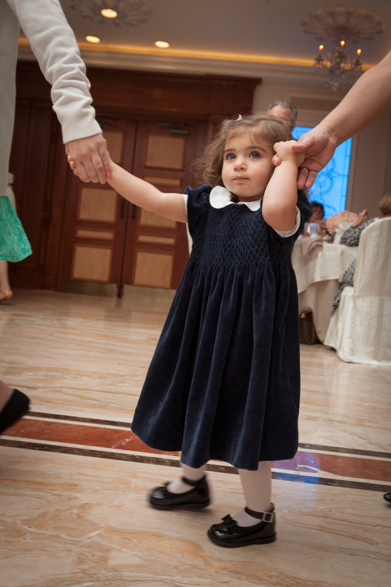 A toddler girl in a navy smocked dress holds adults hands while dancing at a formal reception.