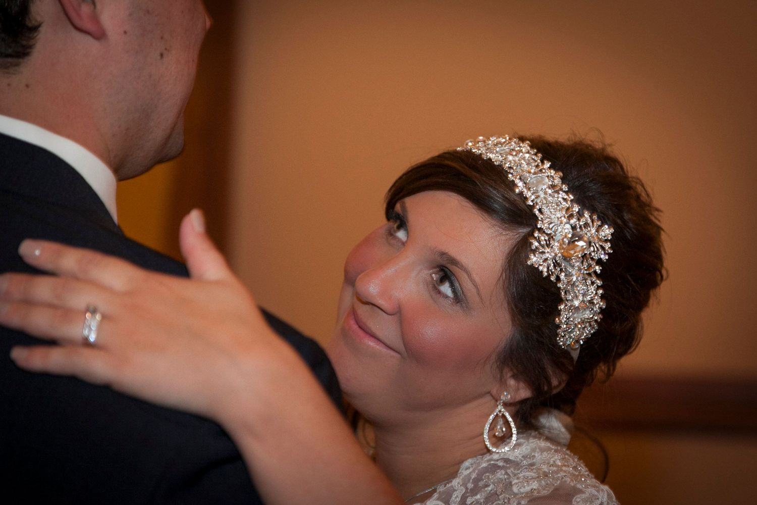 Bride in lace gown with crystal headpiece gazing lovingly at groom during romantic first dance at wedding reception.