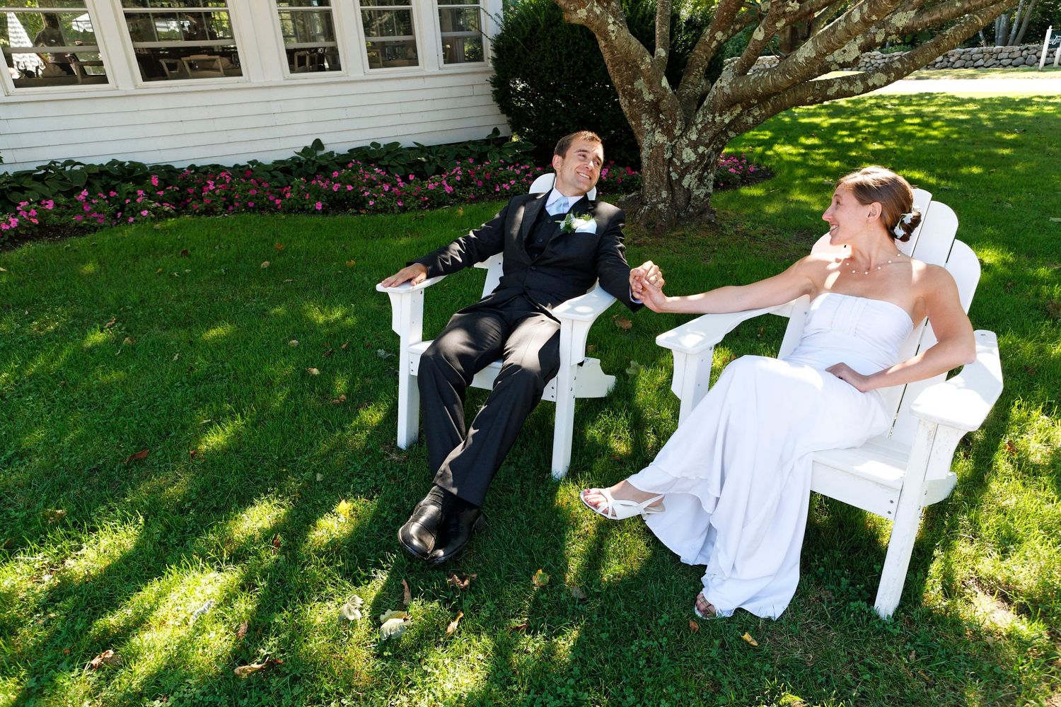 Bride and groom relax in white Adirondack chairs on a lush green lawn at their wedding venue.