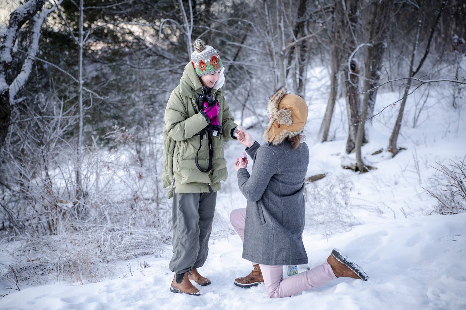 Couple in winter coats exchange rings during snowy outdoor marriage proposal in snowy forest setting.