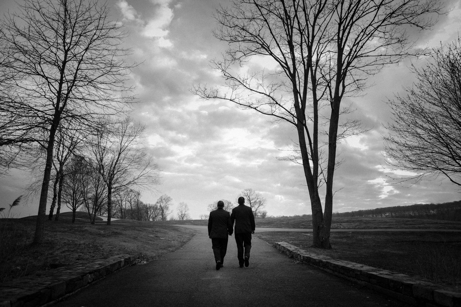 Couple walks hand-in-hand down a tree-lined path under dramatic cloudy skies in black and white.