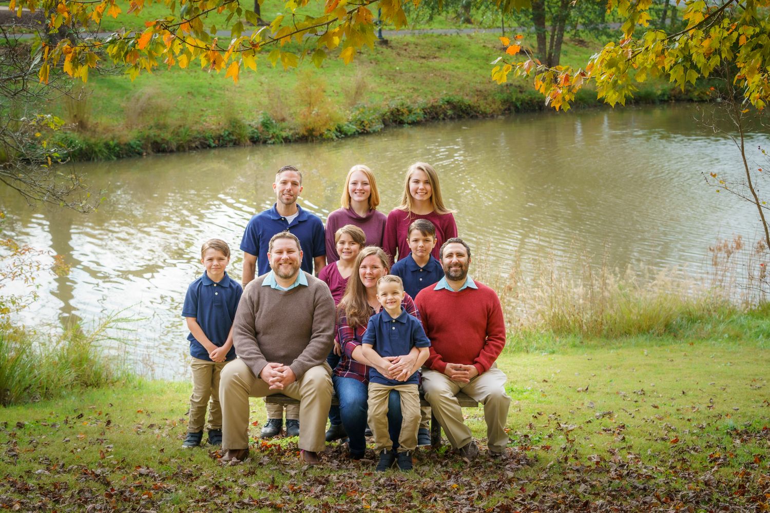 Large family posing outdoors near a pond surrounded by colorful autumn foliage for a fall portrait.