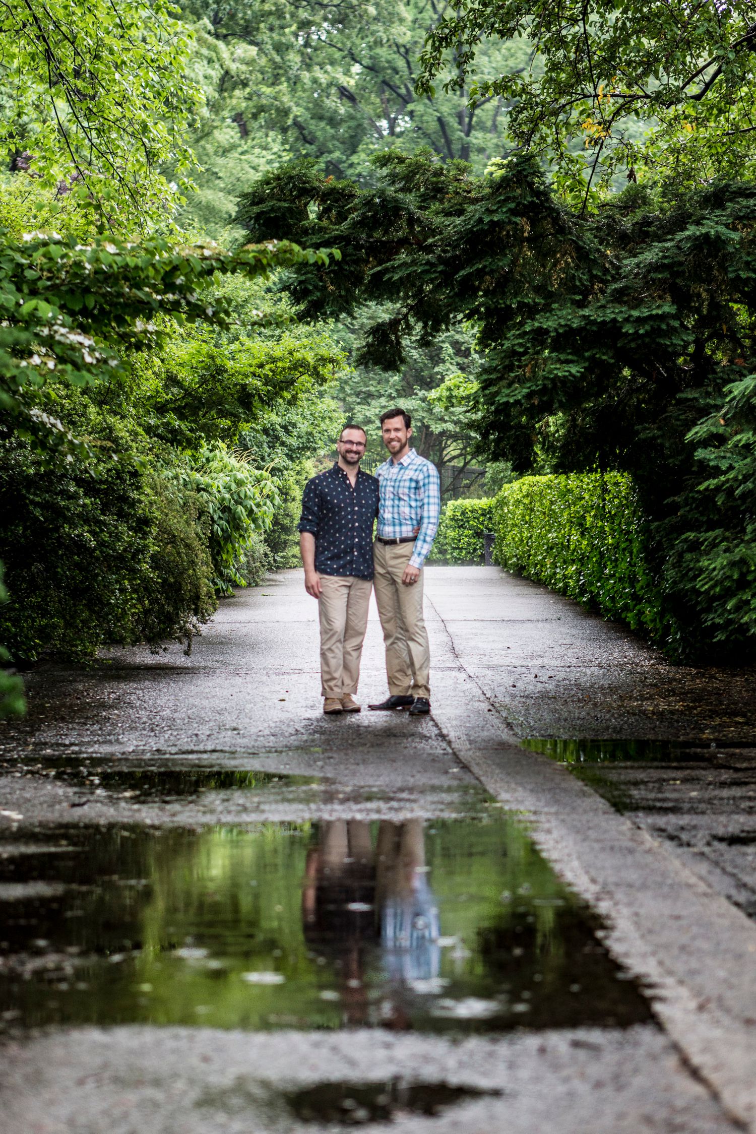 Two men standing on a rain-soaked garden path surrounded by lush green trees, with their reflection visible in a puddle.