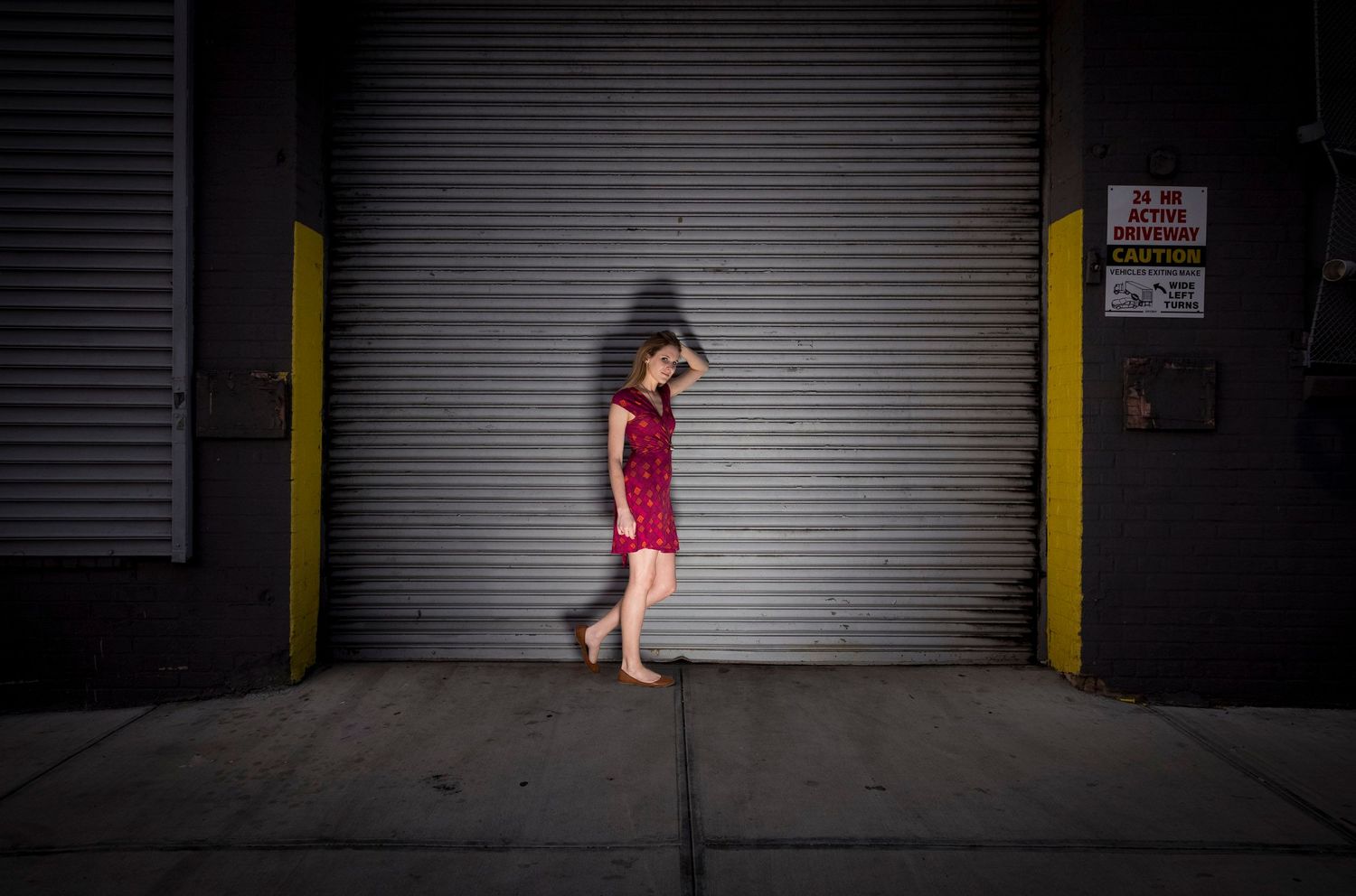 Woman in red dress standing barefoot against a metal shutter door on a city sidewalk.