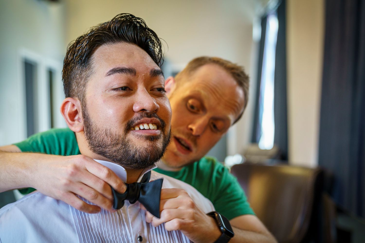 A man in a striped shirt smiles as another man helps him adjust his black bow tie before a wedding.