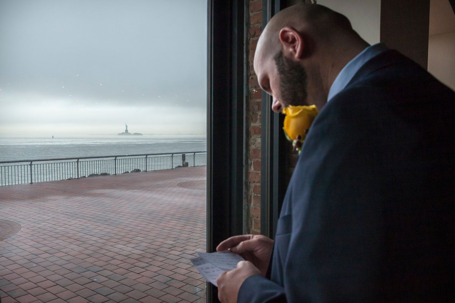 Bald man in suit with yellow boutonniere looks out window at Statue of Liberty on overcast day.