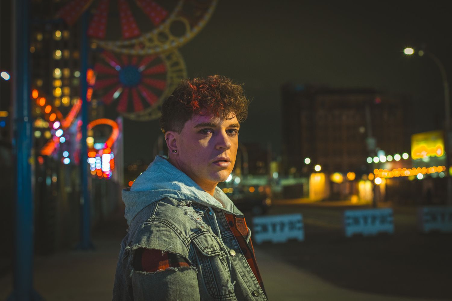 Curly-haired young man in a worn denim jacket looks at the camera at night with a lit amusement park Ferris wheel behind him.