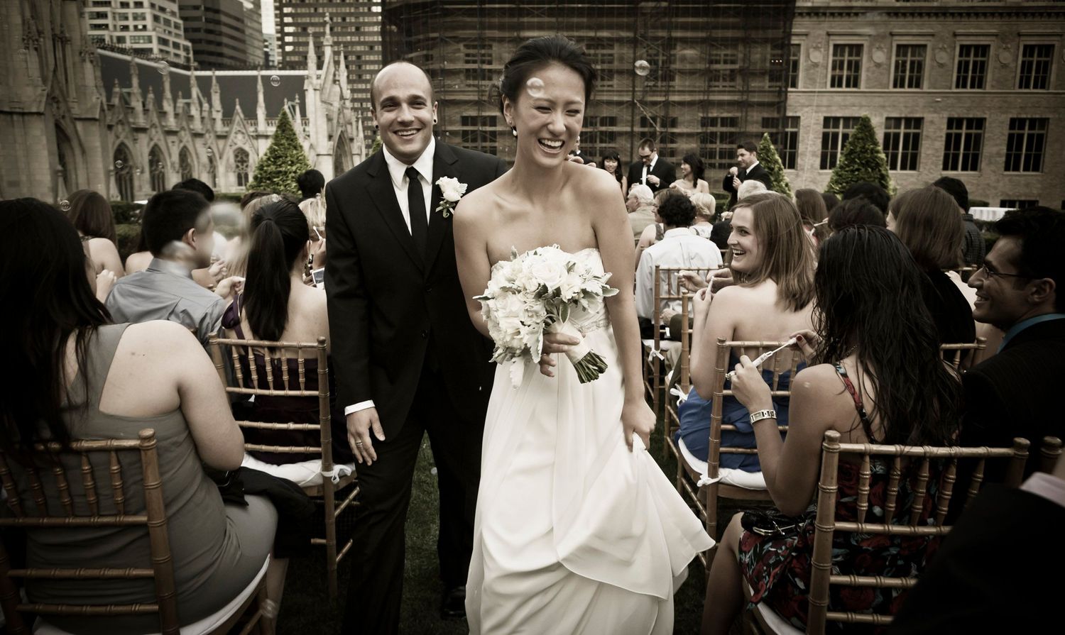 Newlywed couple walks down aisle in sepia-toned photo with wedding guests seated on chiavari chairs.