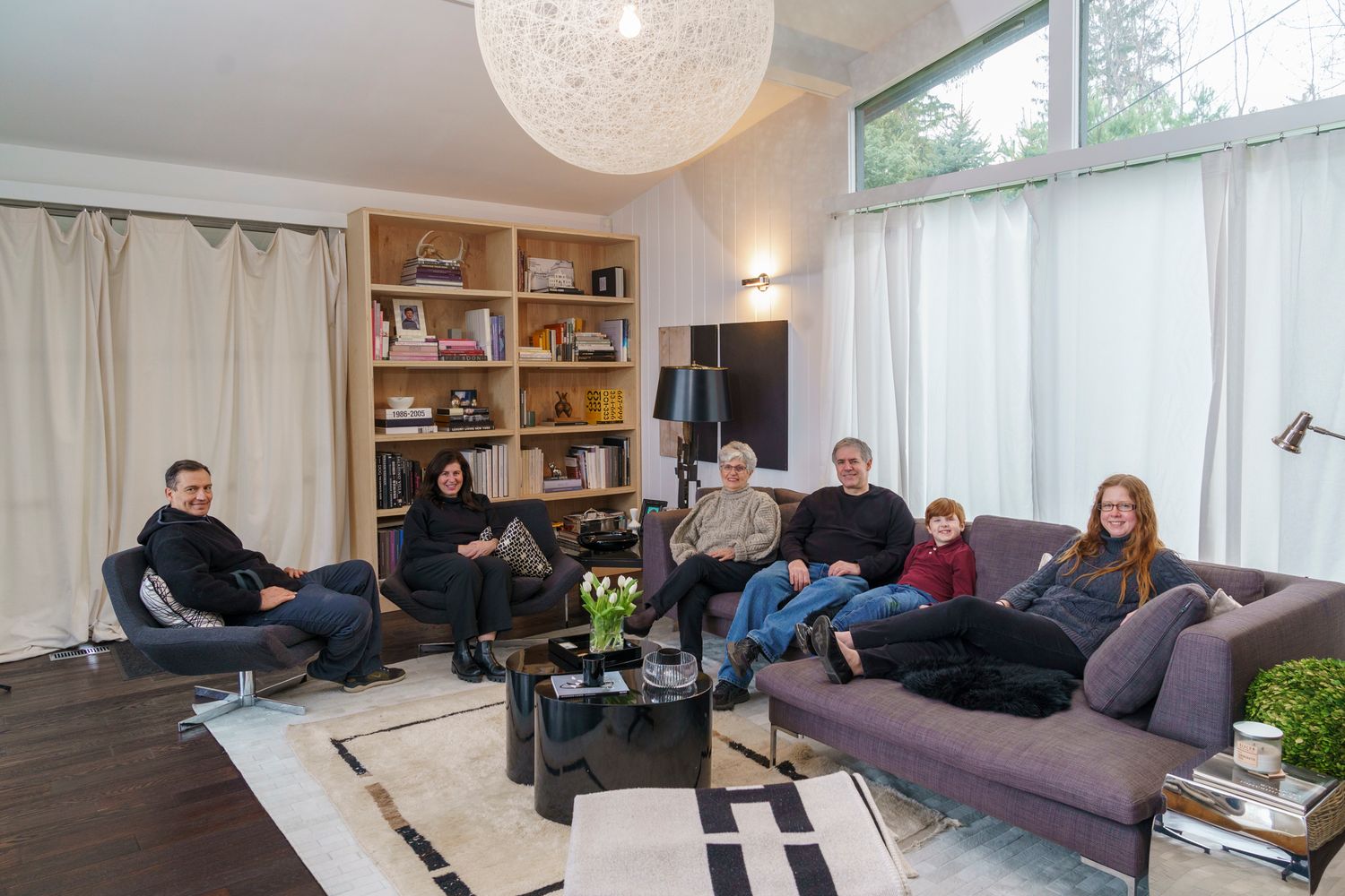 Group of seven people relaxing in a modern living room with bookshelves, large windows, and stylish furniture.