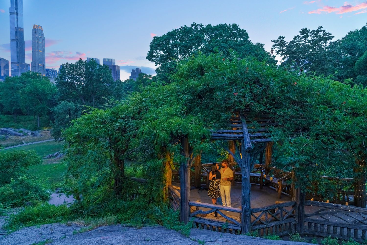 A rustic wooden gazebo nestled among lush green trees in Central Park at dusk with NYC skyline in background.