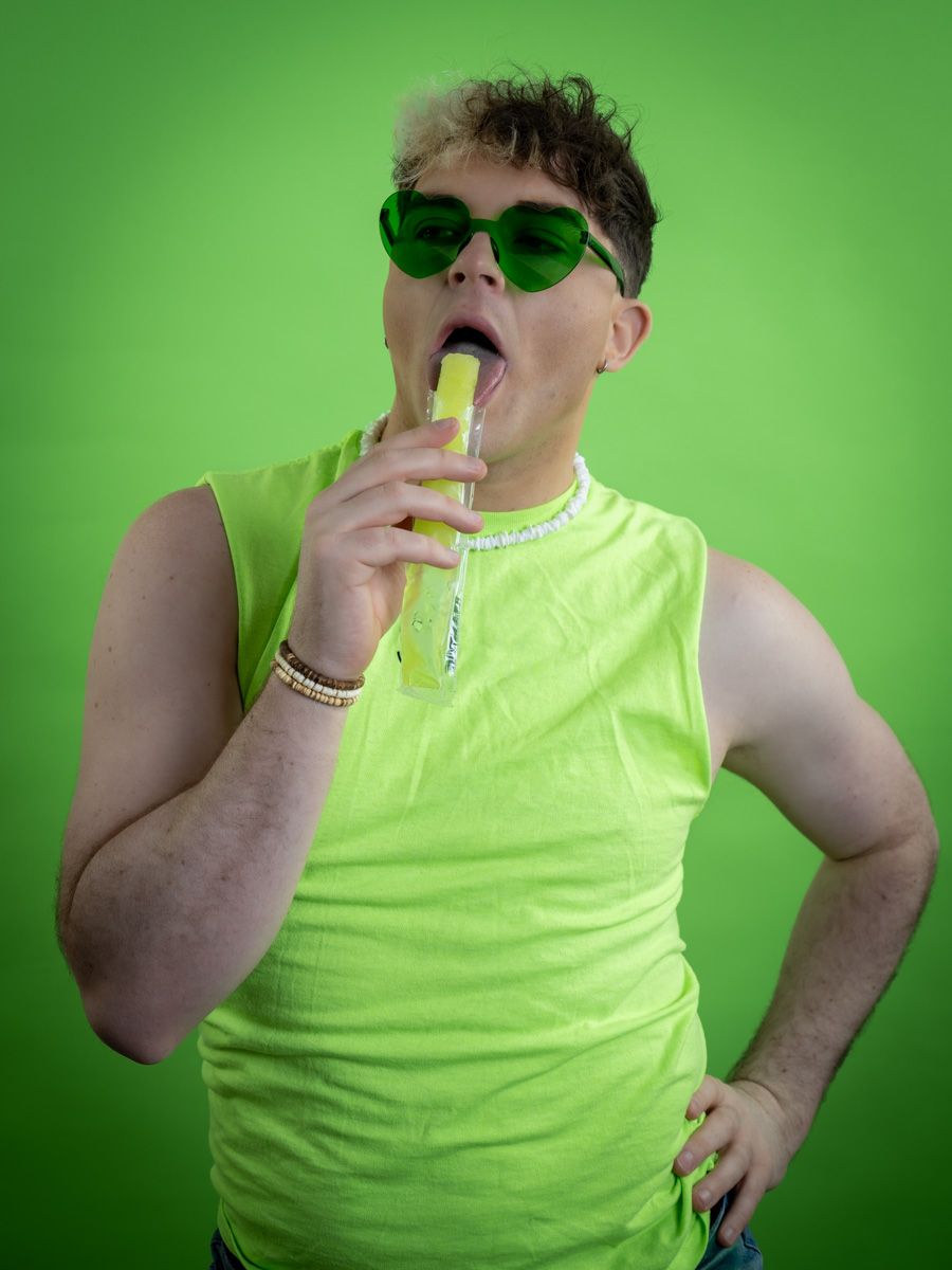 Person in neon green tank top and green sunglasses licking a yellow popsicle against a green background.