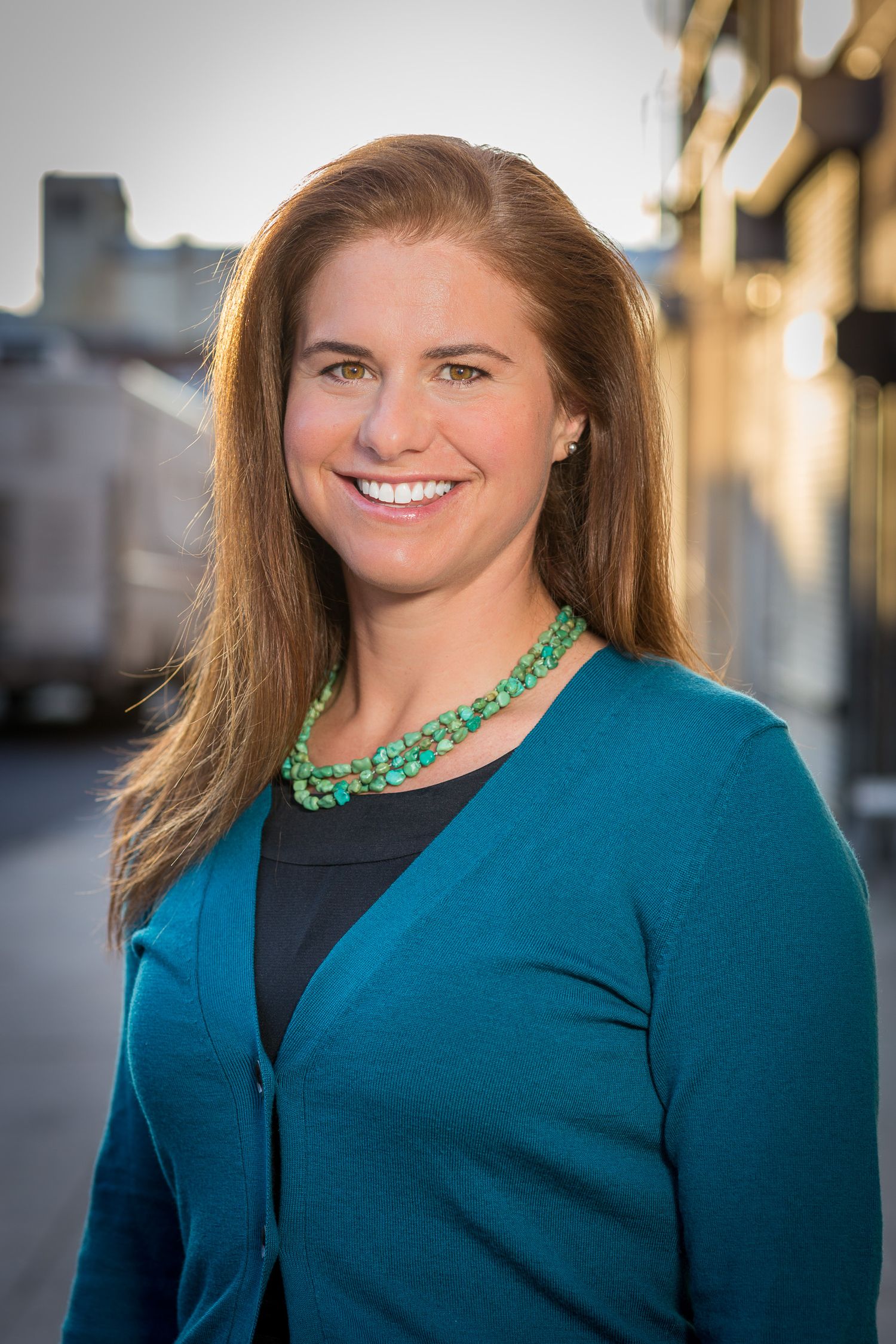 Professional headshot of a smiling woman with auburn hair wearing a teal cardigan and green beaded necklace outdoors.