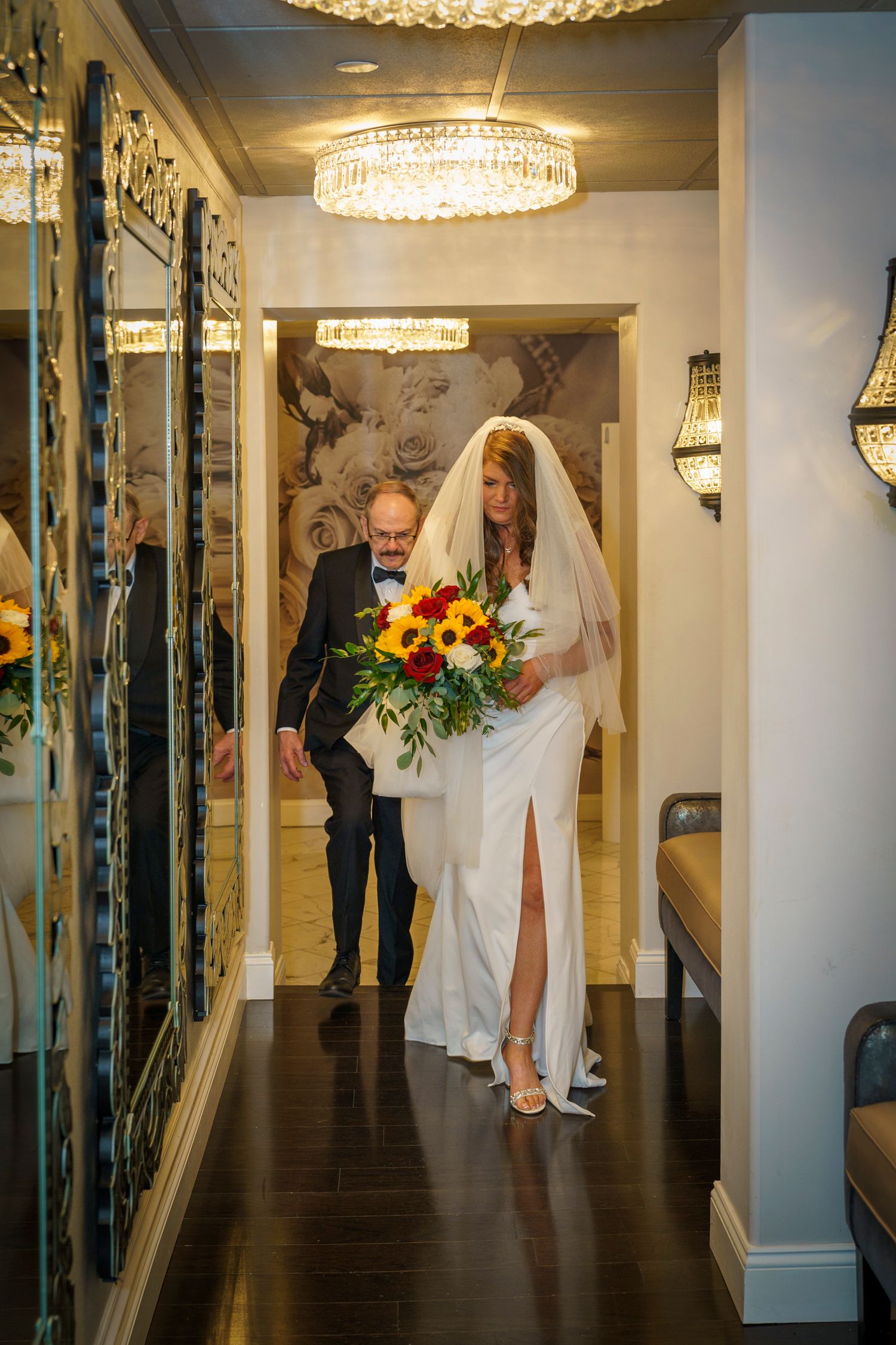 Bride in white gown with veil holding sunflower bouquet walks down elegant hallway with escort under crystal chandelier.