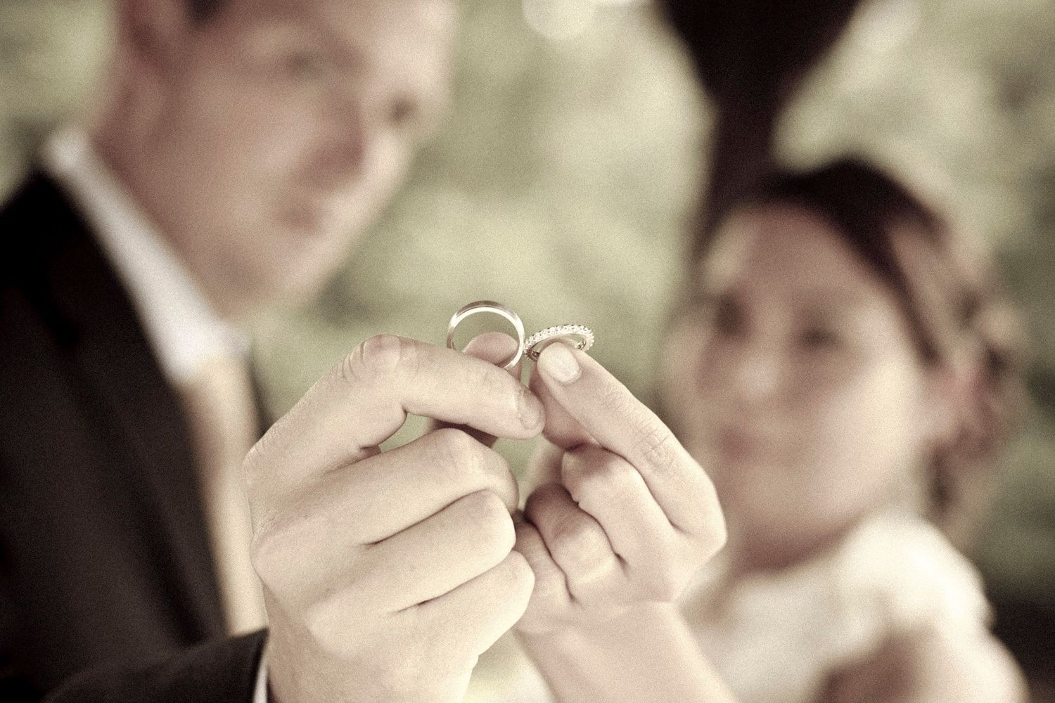 Newlywed couple holding interlocked wedding rings together in a soft sepia-toned romantic photo.