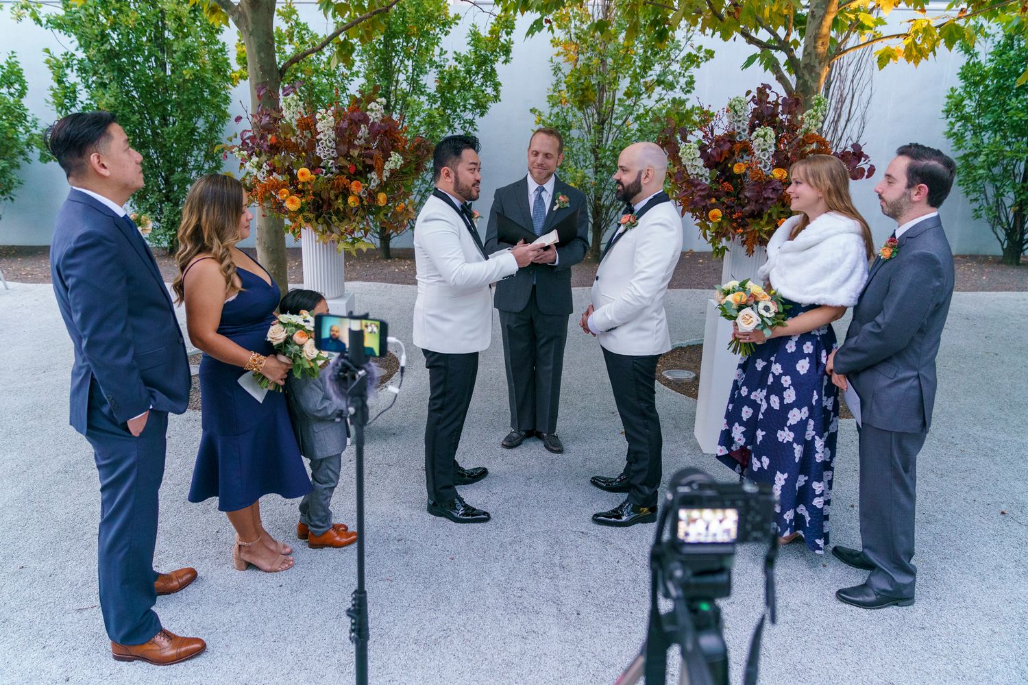 Two grooms exchange vows outdoors at a fall wedding ceremony with guests and floral arrangements.