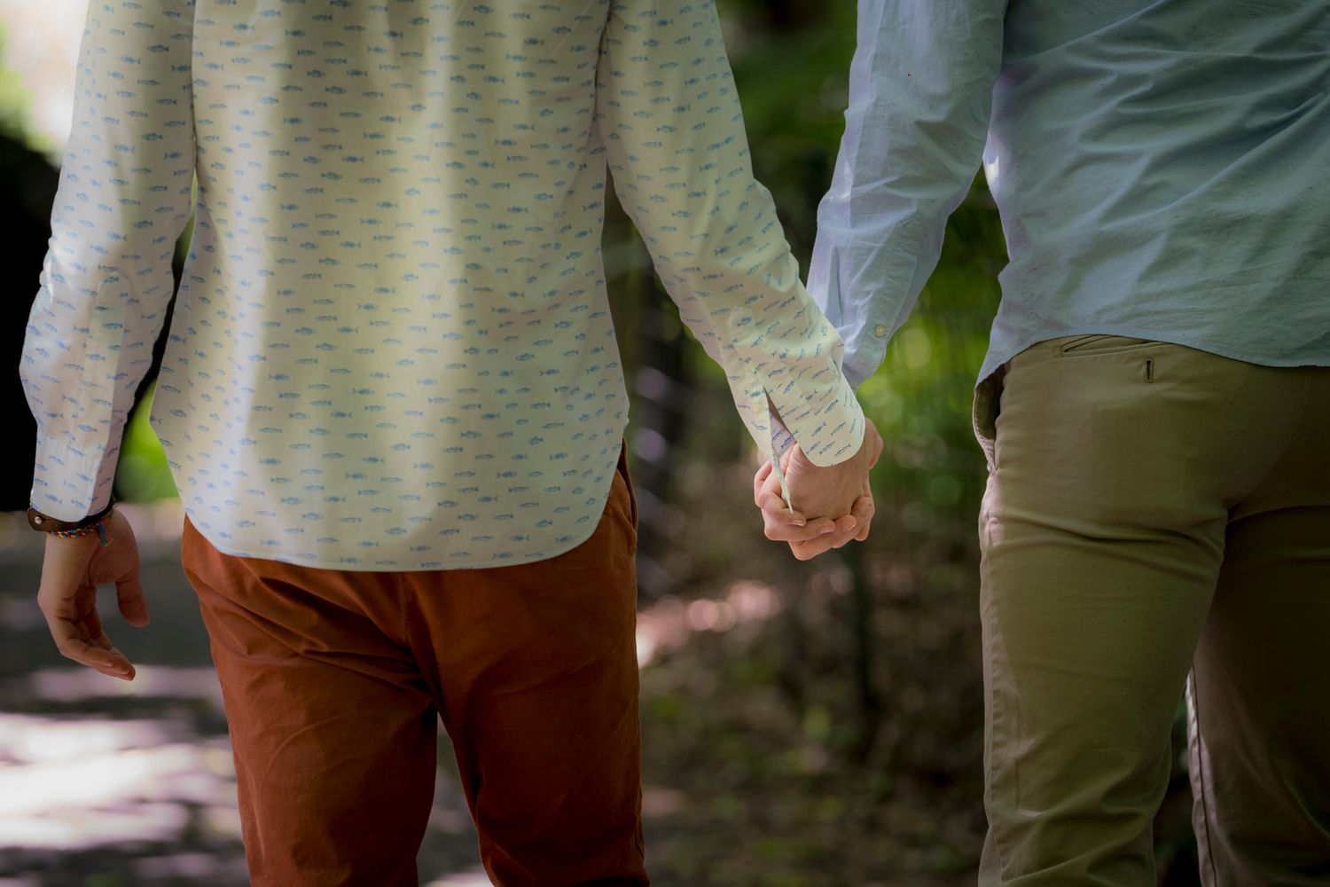 Couple holding hands while walking outdoors on a sunny day, wearing casual shirts and khaki and rust-colored pants.