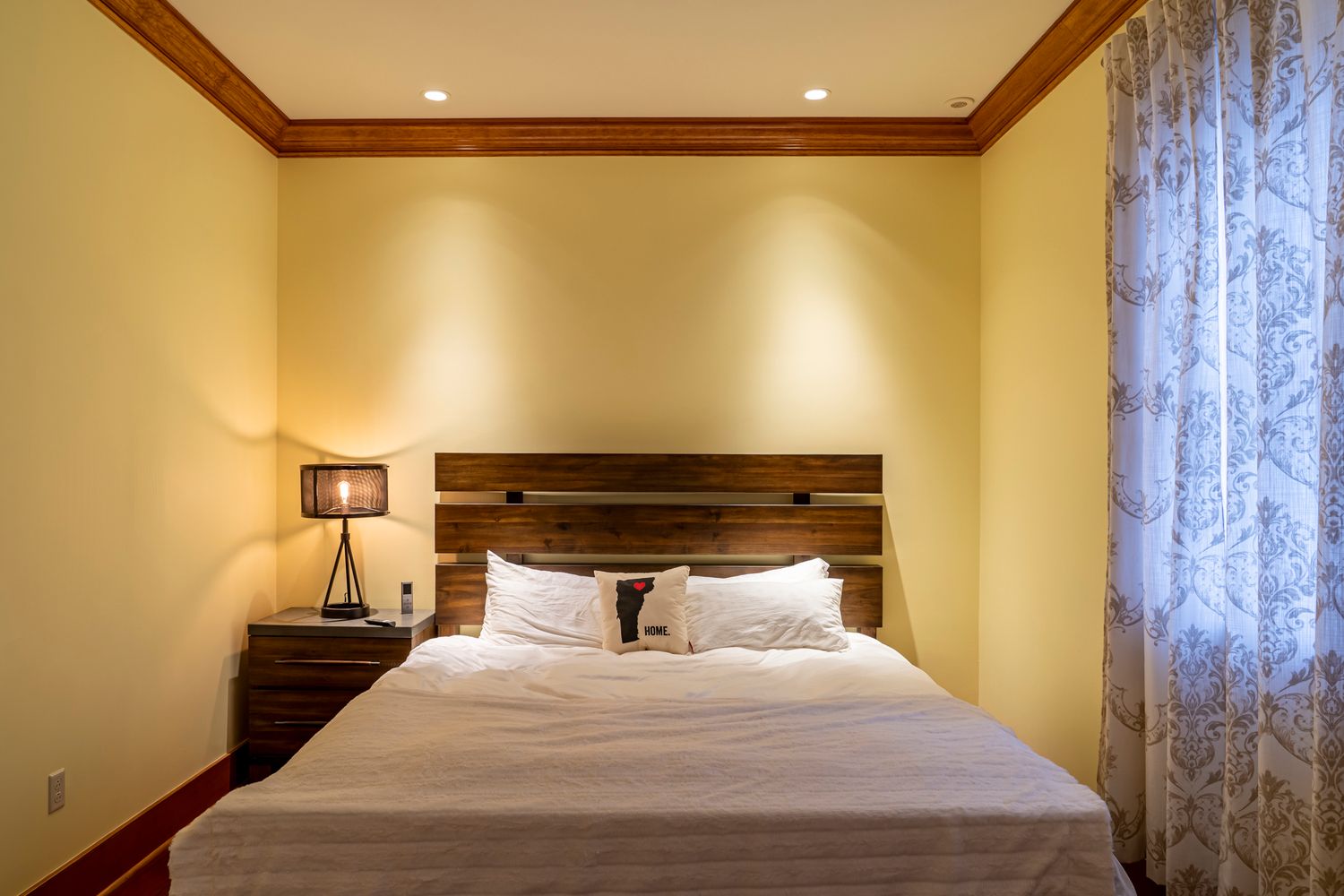 Cozy hotel bedroom with a queen bed, white linens, wooden headboard, nightstand lamp, and soft yellow walls.
