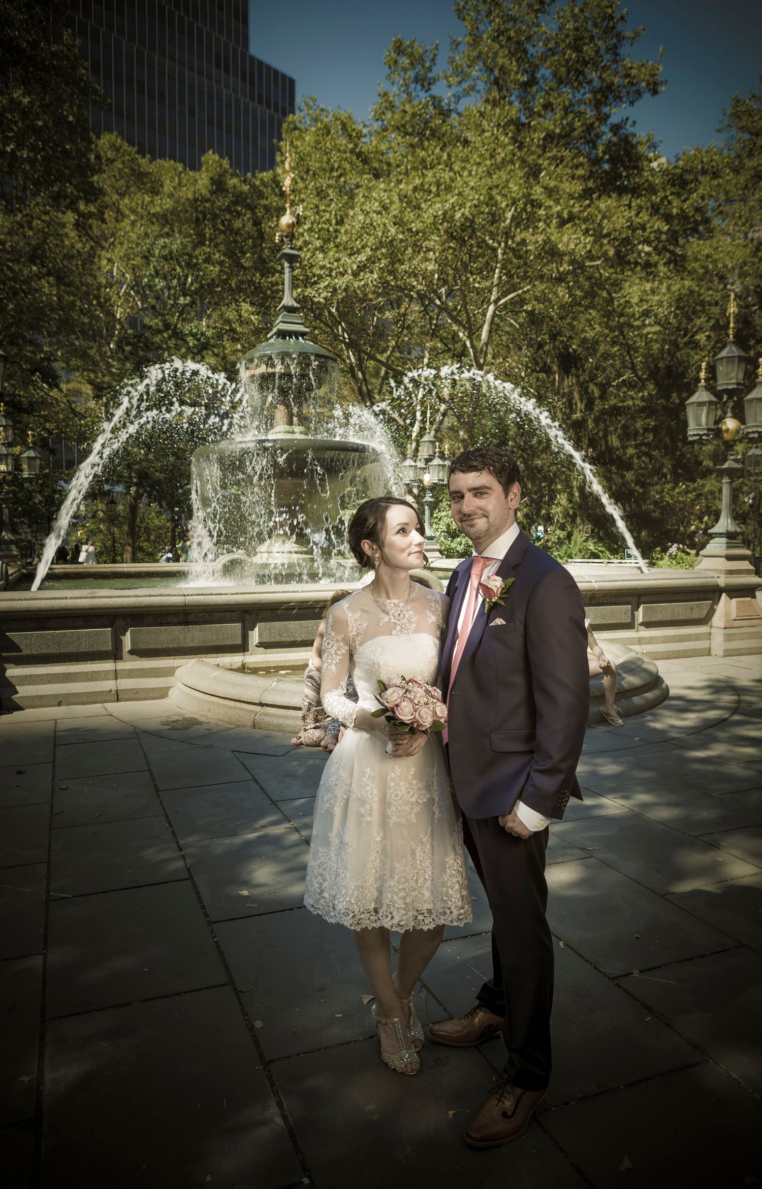 A bride and groom pose romantically in front of a decorative fountain in a park on their wedding day.
