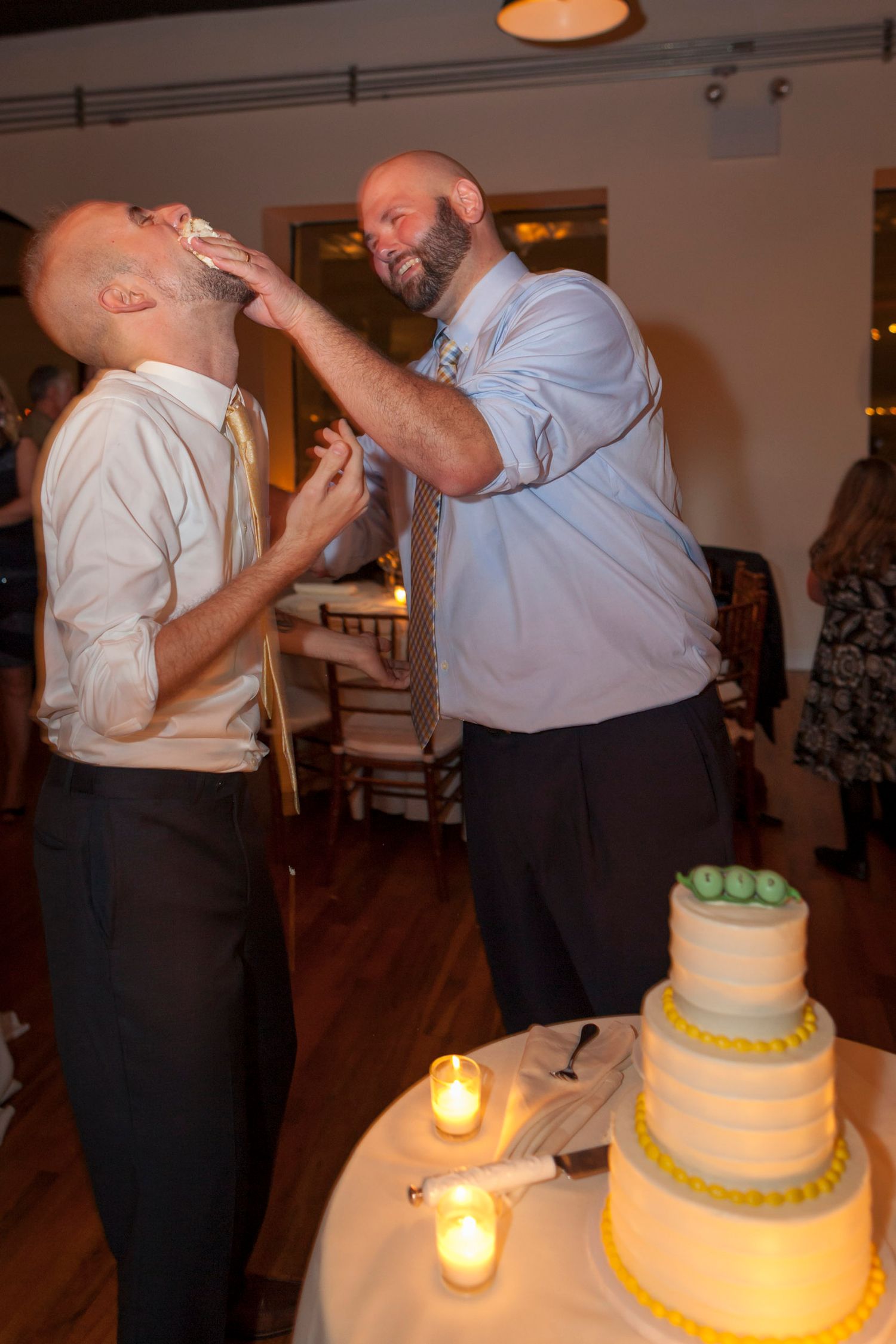 Two grooms share a fun cake smashing moment at their wedding reception near a white tiered cake.