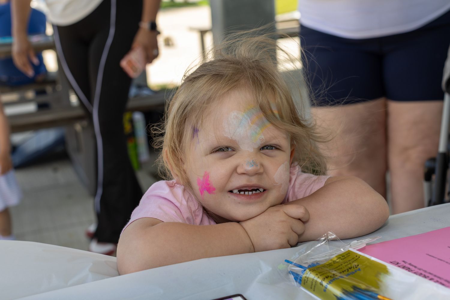 Smiling toddler girl with colorful face paint resting her chin on arms at an outdoor community event.